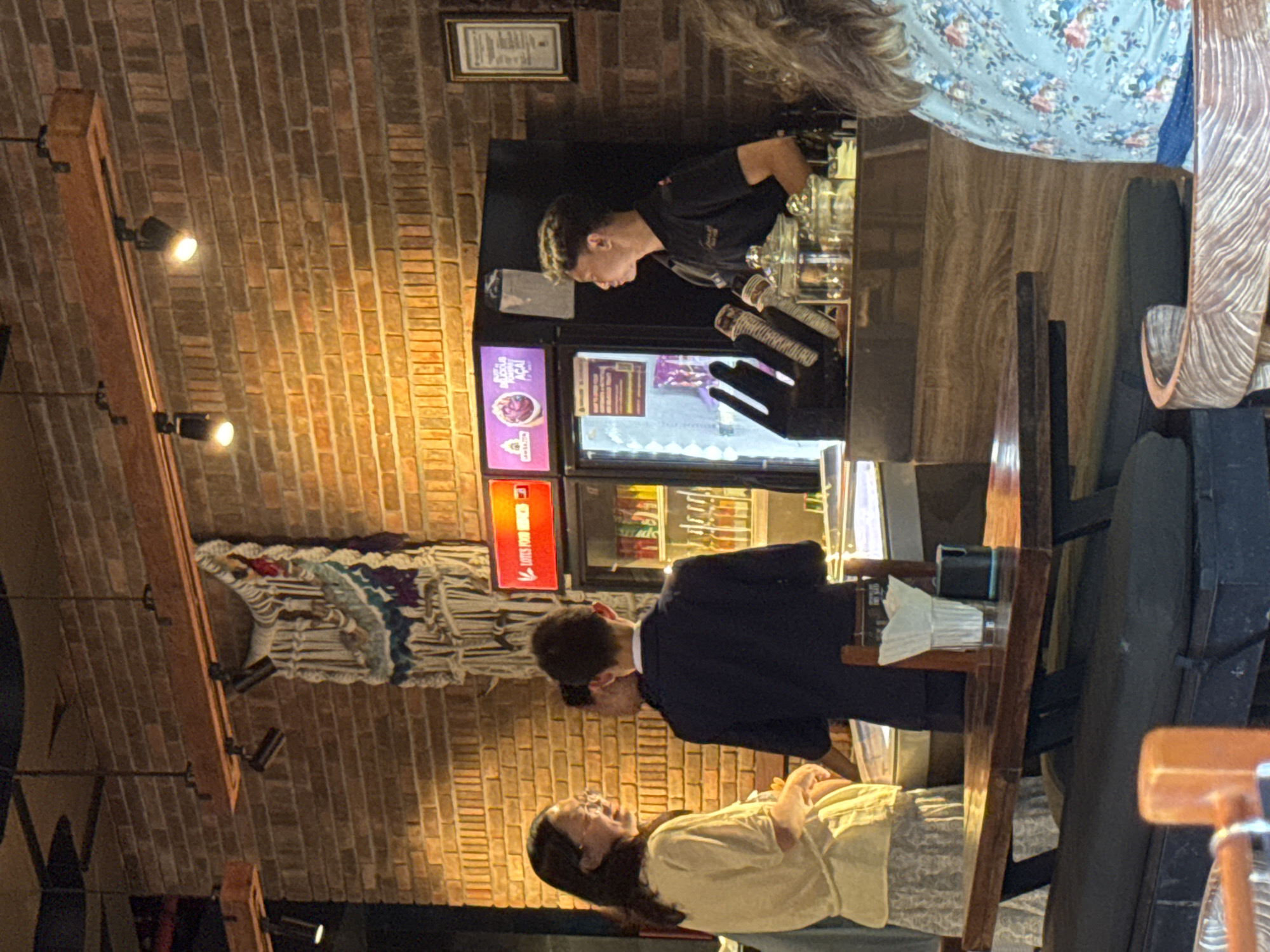 In Kuta Selatan, Indonesia, at a dimly lit establishment with a prominent brick wall, three individuals are observed. A woman with glasses and a light-colored top stands to the left, interacting with a person in a dark suit who is facing away from the camera. To the right, a man in a black polo shirt stands behind a counter, facing a glass-fronted refrigerator filled with colorful drinks. He appears to be attending to customers or preparing orders. The refrigerator displays illuminated signs, one red with white text and another purple with white text. The counter area is equipped with various containers, possibly for ingredients or condiments, and a napkin dispenser. In the foreground, a wooden table with a dark upholstered bench is visible, suggesting a seating area within the establishment. The overall atmosphere is that of a casual dining or beverage service venue.