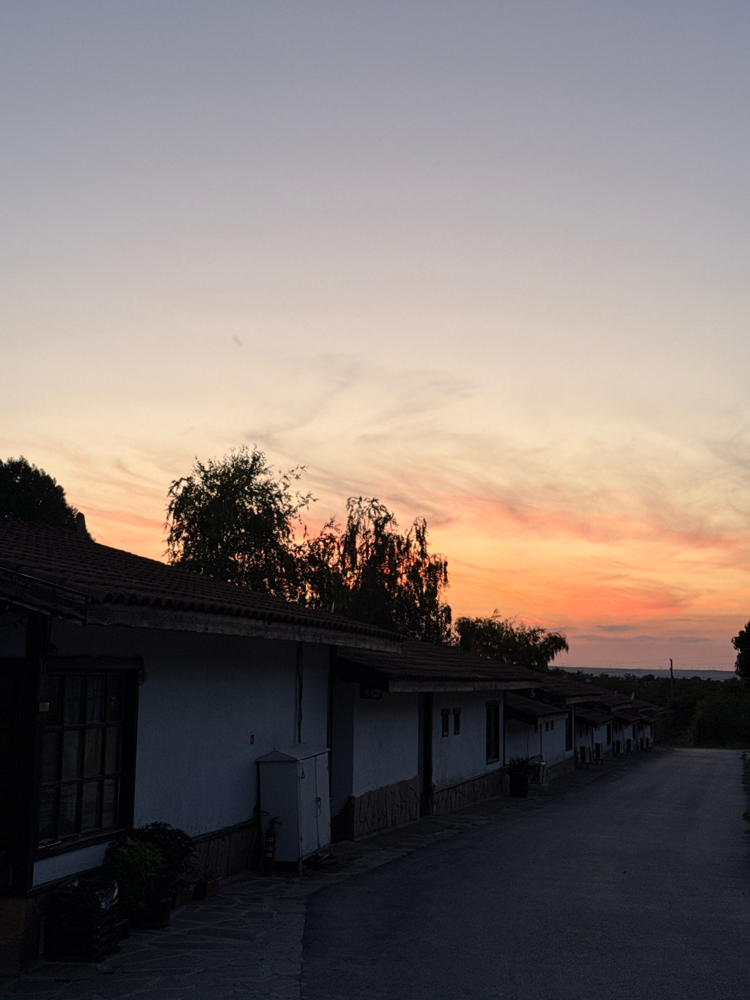 The image depicts a tranquil scene in Chernevo, Bulgaria, during a sunset. The sky displays a gradient of soft purples and oranges, with wispy clouds suggesting the fading light. In the foreground, a row of white, single-story buildings with dark tile roofs lines a paved road. These structures appear to be houses or small bungalows, with some featuring visible windows and air conditioning units. Dark trees and foliage are silhouetted against the colorful sky, adding depth to the landscape. The road itself is empty, contributing to the peaceful atmosphere of the evening. A small, grey electrical box and a red fire extinguisher are visible next to one of the buildings. The overall impression is one of quiet solitude and natural beauty as day transitions into night.