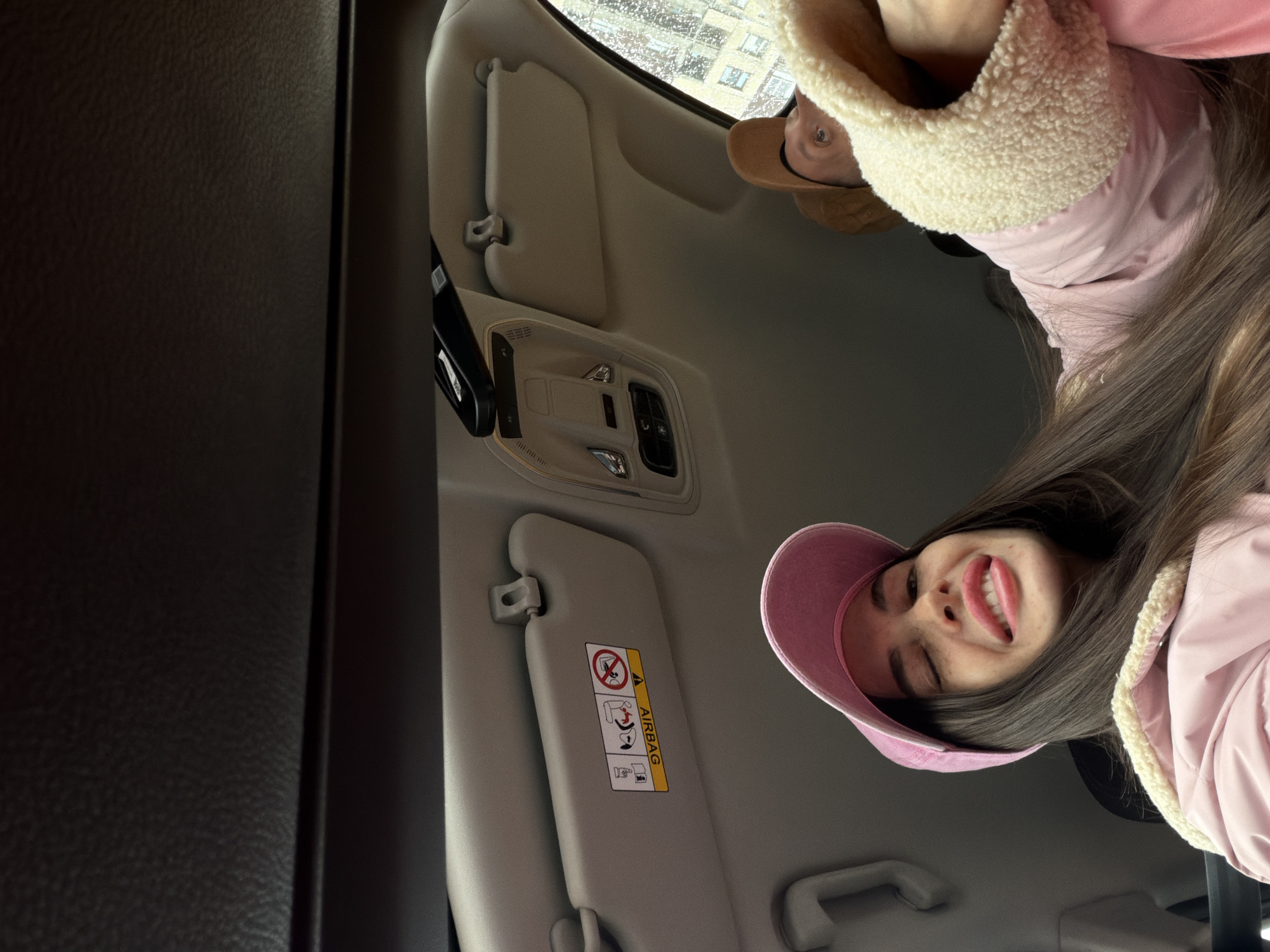 A close-up shot from inside a vehicle shows two people, with the primary focus on a young woman in a pink baseball cap and jacket with a furry collar. She is smiling with her eyes partially closed. Another person wearing a tan hat and jacket with a furry collar is visible behind and above her. The interior of the vehicle is visible, including the ceiling, sun visors, and a black car door panel. The sun visor has a warning sticker about airbags. The exterior visible through the car window suggests a wet, urban environment with buildings. Based on the architectural style visible through the window and the presence of Cyrillic text in the airbag warning, it is plausible the location is Saint Petersburg, Russia.