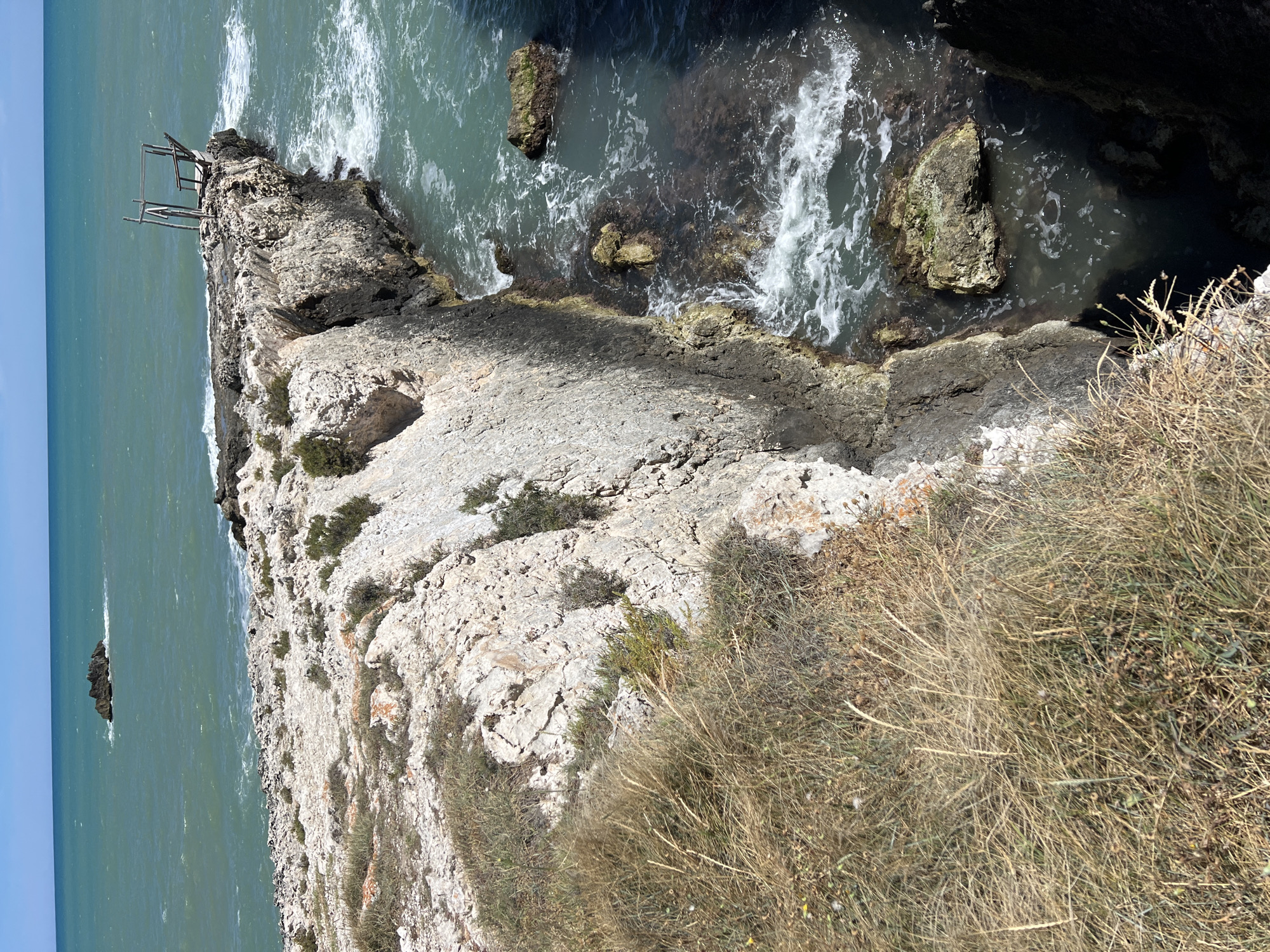 The image shows a rocky coastline with a wooden structure on a cliff edge, overlooking the Adriatic Sea. The sea is a vibrant turquoise, with white waves crashing against the rocks. There is a small rock formation in the distance. The cliff is covered in sparse vegetation, including dry grass and small bushes. The wooden structure appears to be a fishing platform or observation point, with a simple wooden frame and a deck. The scene suggests a natural, rugged landscape typical of the Gargano Peninsula in Italy. The location is likely Peschici, a coastal town known for its dramatic cliffs and clear waters. There are no people or animals visible in the image.