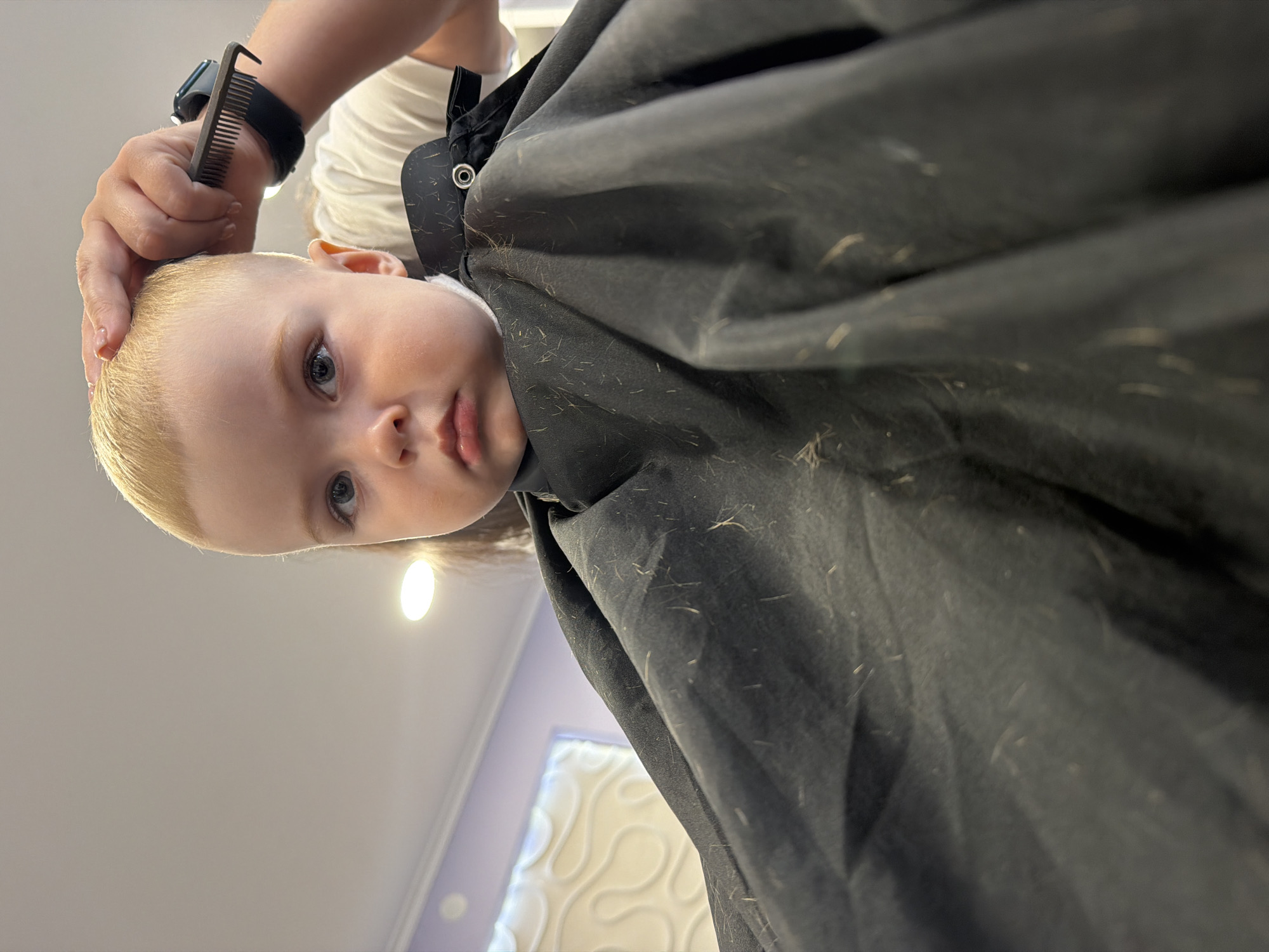 A toddler with blond hair is getting a haircut. A person is holding a comb near the toddler's head. The toddler is wearing a black barber cape with stray hairs visible. The location is likely a barber shop in Votkinsk, Russia. A light fixture and decorative wall panel are visible in the background.