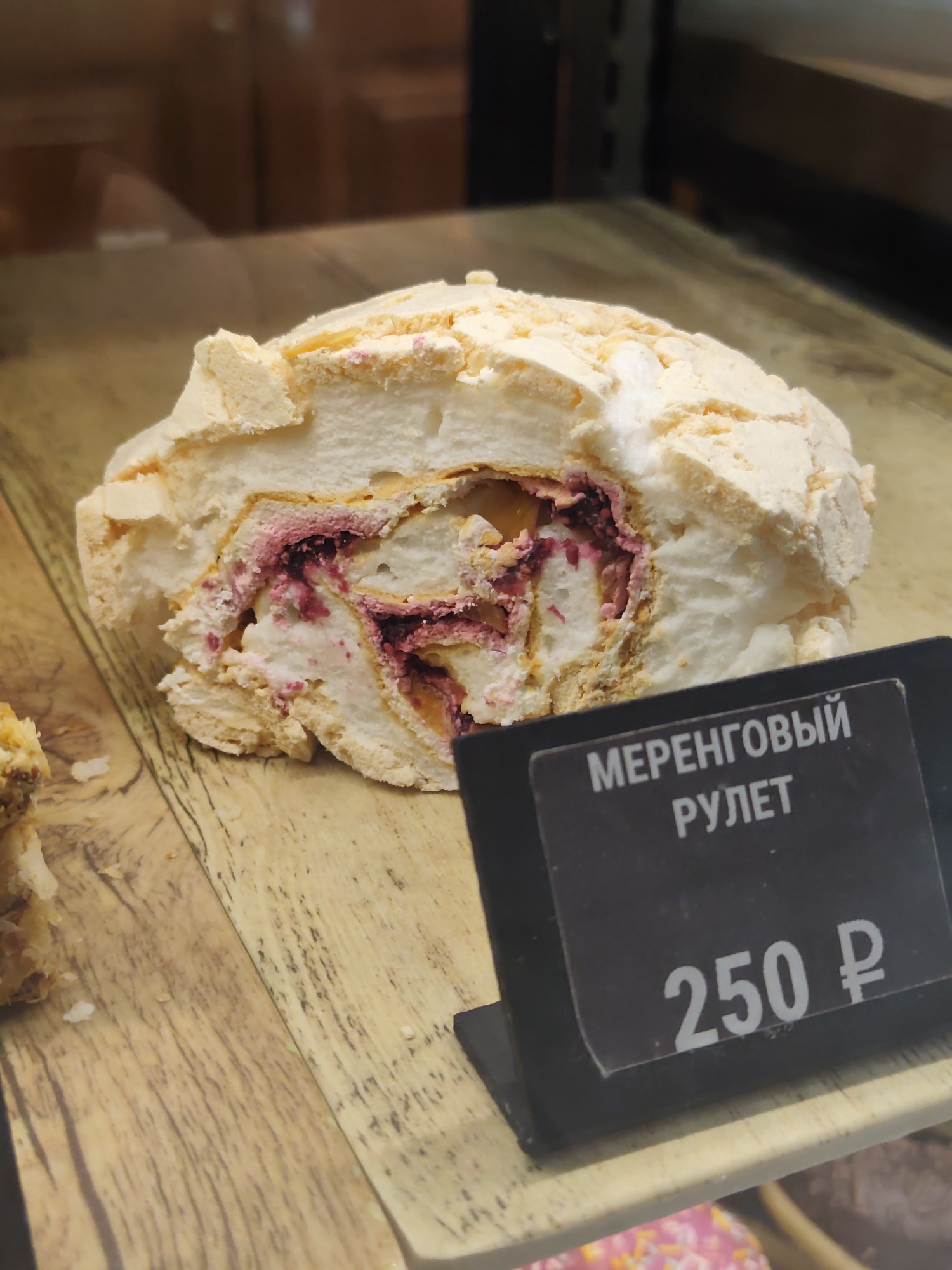 A close-up shot showcases a slice of a meringue roll on a wooden surface. The roll, light in color with a crisp meringue exterior, is filled with a creamy white filling and a vibrant pinkish-purple berry compote. A small black sign in the foreground displays Russian text, "МЕРЕНГОВЫЙ РУЛЕТ", meaning "Meringue Roll", and the price "250 ₽". The background is blurred, showing a display case and a hint of other baked goods. The scene is likely from a bakery or cafe in Sochi, Russia.