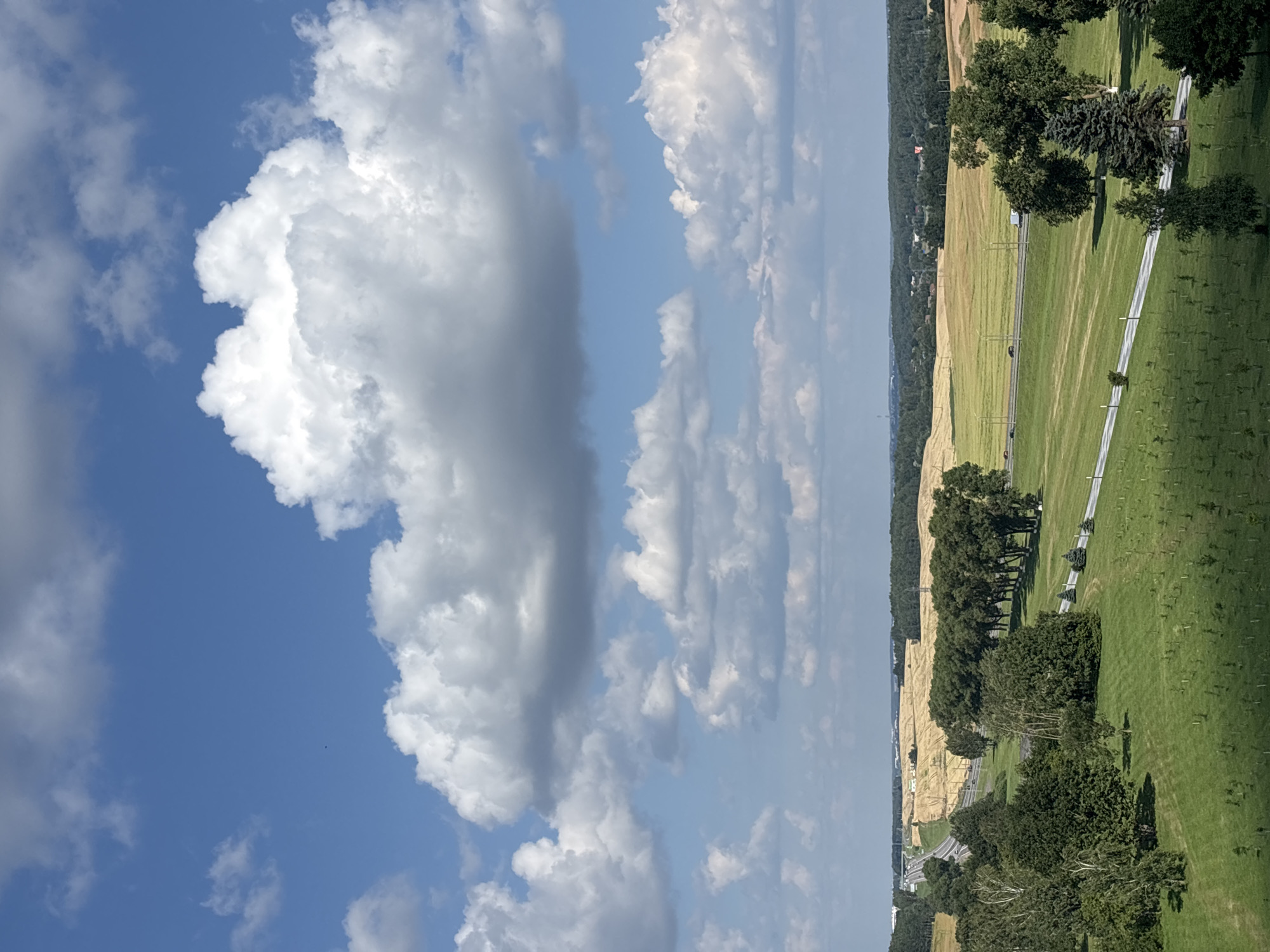 This is an aerial, full-frame, eye-level, wide shot of a rural landscape in the city of Ozyaritska-Slabadski rural council, Belarus. The scene is dominated by a blue sky with fluffy white and gray clouds. Below the sky, the landscape features rolling green fields, a highway with cars and trucks, and a line of mature trees. In the foreground, a neatly mowed green lawn is dotted with small saplings, suggesting a recently planted area or a young orchard. The middle ground showcases a paved road winding through a green field, bordered by a row of tall, dense trees. Further back, a golden-brown field stretches towards a distant treeline, with power lines visible in the distance. The overall impression is one of a serene, agricultural countryside under a bright, partly cloudy sky.
