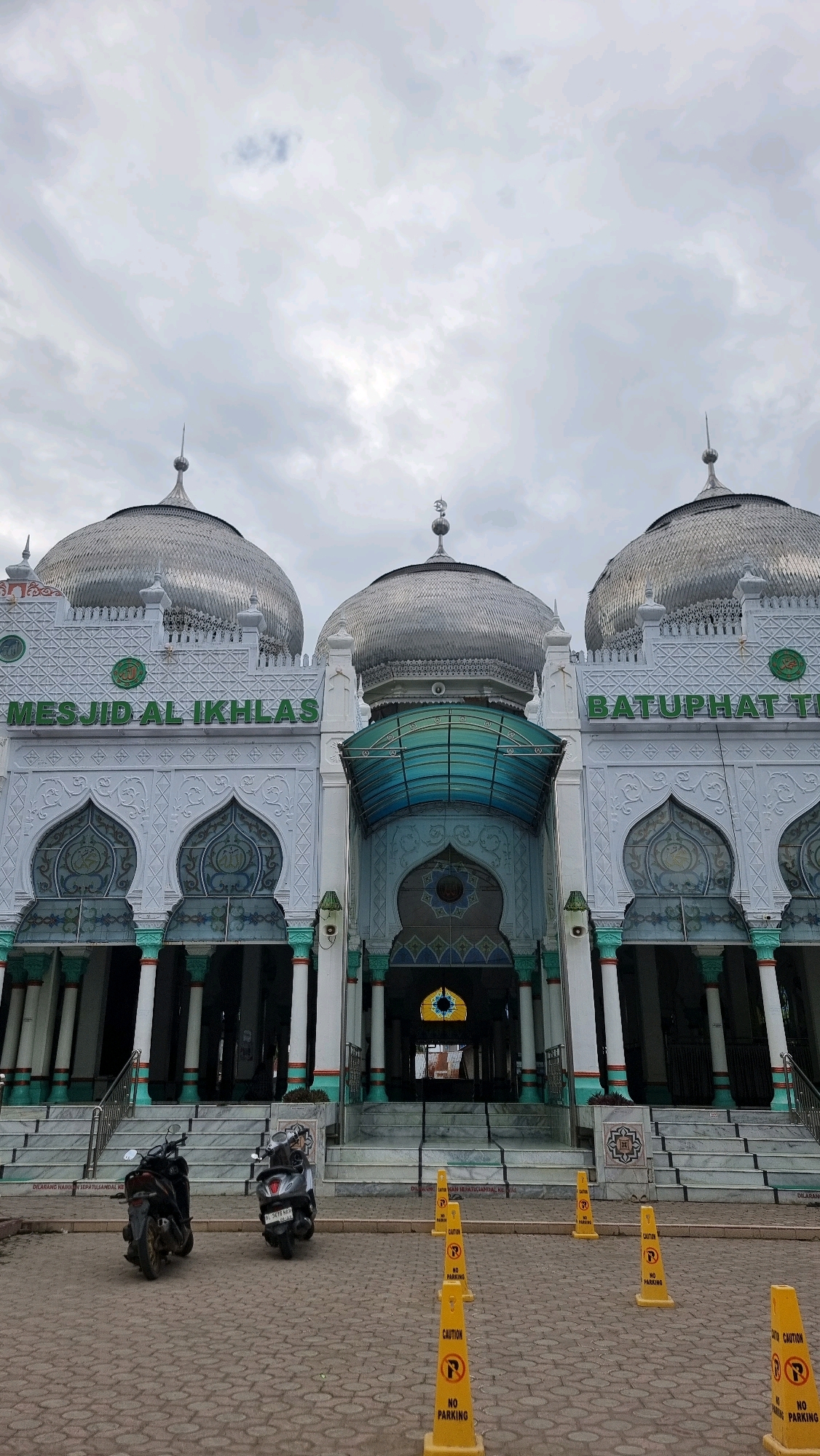 The image shows the exterior of the Masjid Al Ikhlas in the city of Lhokseumawe, Indonesia. The mosque features a prominent facade with large, silver-colored domes, intricate architectural details, and green lettering that spells out "MESJID AL IKHLAS" and "BATUPHAT TH". The entrance is flanked by tall columns and ornate archways. In front of the mosque, on a paved courtyard, are two parked motorcycles and several yellow "NO PARKING" cones. The sky above is overcast with clouds.