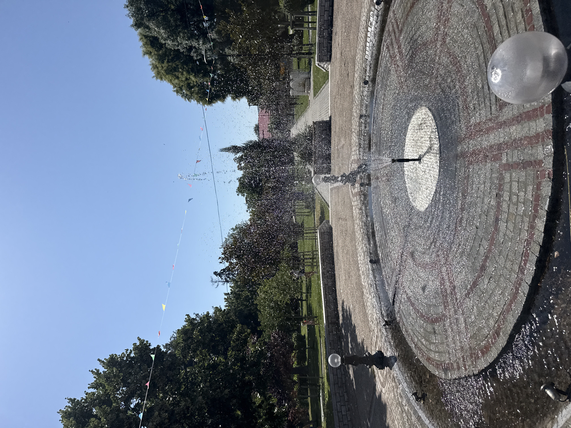 The image depicts a fountain in a public park in Strelcha, Bulgaria. The fountain, circular in shape and paved with grey and red stones, is actively spraying water. The water droplets catch the sunlight, creating a shimmering effect.

Surrounding the fountain are paved pathways and green lawns with scattered trees. In the background, a pathway leads towards buildings and more trees, suggesting a well-maintained urban park area. Colorful triangular flags are strung across the sky, adding a festive touch to the scene. The sky is clear and blue, indicating fair weather.

There are a few people visible in the distance, appearing to be children playing on the grass, their forms blurred by distance. A decorative lamp post stands to the left of the fountain, and another translucent spherical light fixture is visible in the foreground on the right. The overall impression is of a bright, sunny day in a pleasant, recreational space within the city.