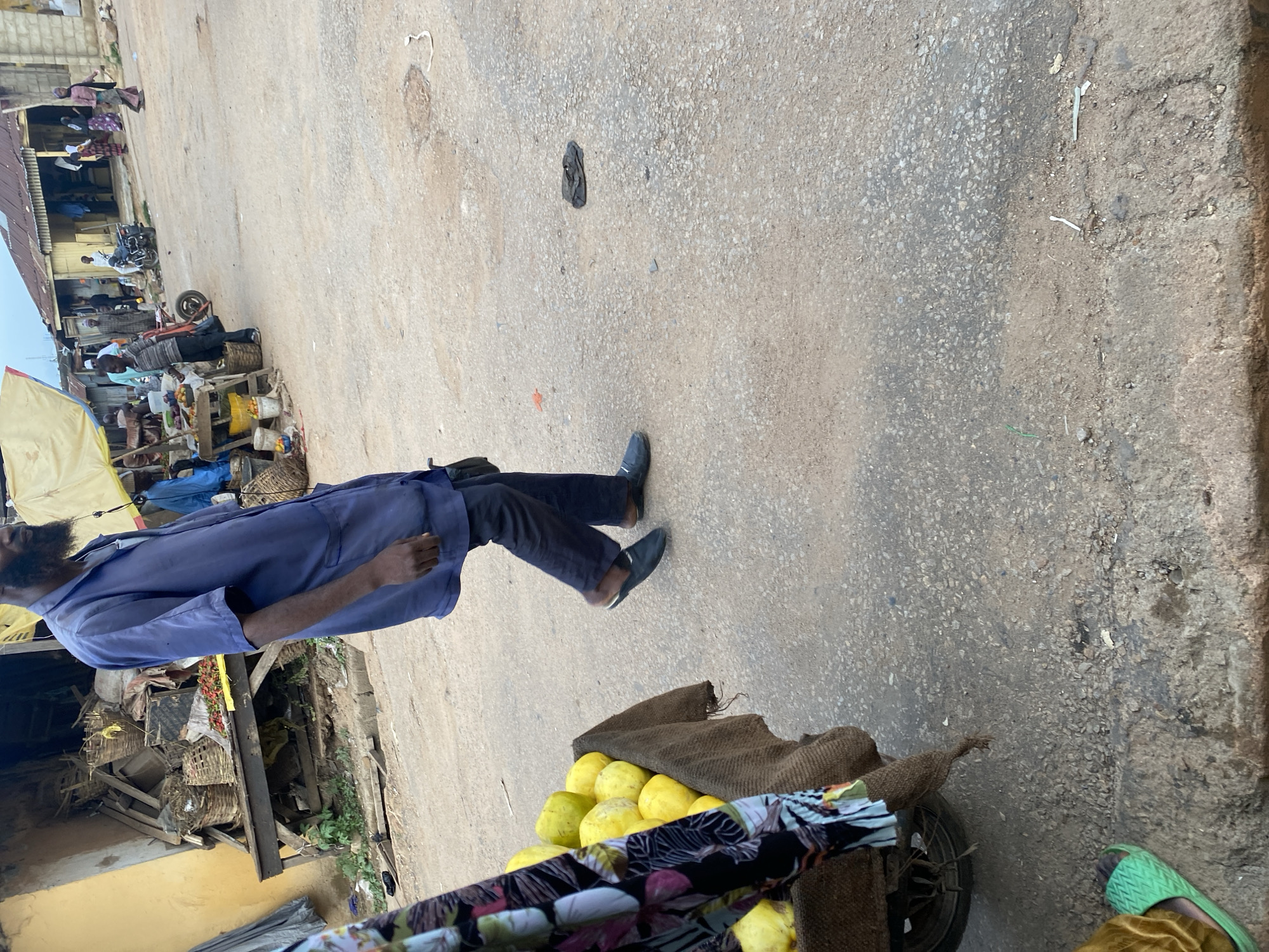 The image depicts a street scene in Jos, Nigeria, likely a market area. In the foreground, a person wearing a blue shirt and dark pants walks away from the camera. To the left, a cart filled with yellow fruits, possibly mangoes or papayas, is partially visible. Behind the cart, another person's legs are visible, wearing a green sandal and golden fabric. In the background, market stalls with various goods, including baskets and produce, are set up. Several other people are present in the background, some walking and others near the stalls. The ground is a rough, paved surface with some debris. The overall atmosphere suggests a busy, open-air market environment.