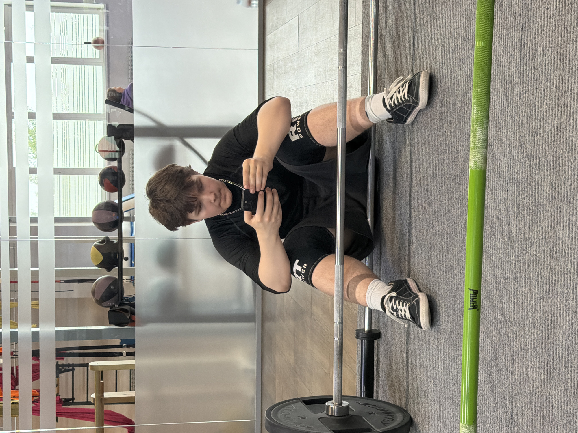 A young man is sitting cross-legged on a grey carpeted floor inside what appears to be a gymnasium or fitness center. He is wearing black shorts with the word "POWER" printed on the leg, a black t-shirt, white socks, and black sneakers with white laces. He is holding a smartphone in his hands and appears to be taking a selfie or a photo. A barbell with a weight plate is placed in front of him. To his right is a green-colored barbell. Behind him is a wall with a mirror reflecting exercise equipment, including medicine balls on a rack, and a window with vertical blinds, offering a partial view of the outside. The location is in the city of Chaikovsky, Russia.