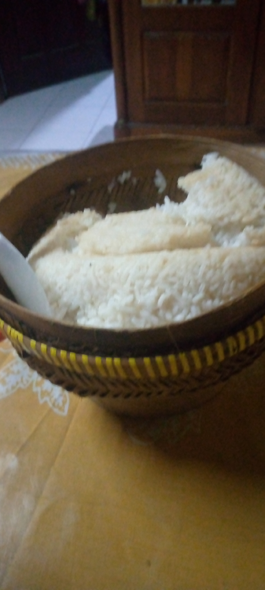 In the city of Sumberwono, Indonesia, a close-up shot focuses on a woven basket filled with white rice. The basket has a woven pattern with yellow accents along the rim. A white spoon is partially visible on the left side of the basket, resting on the rice. The rice appears to be cooked and is piled high in the basket, with some unevenness on the surface. The background is out of focus, showing a dark wooden furniture piece and a lighter colored surface, possibly a floor. The overall scene suggests a simple domestic setting where food is being presented.