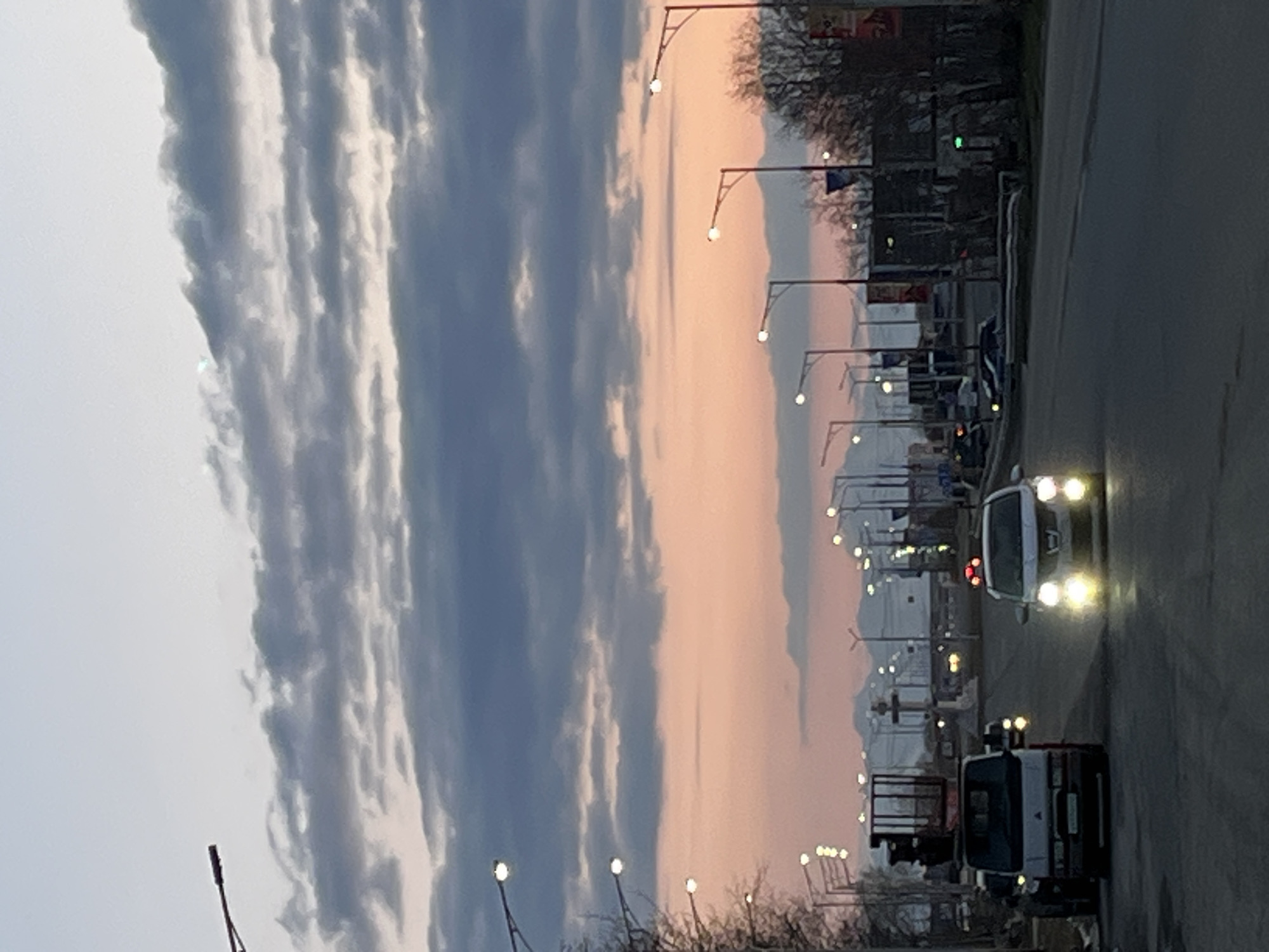 The image depicts a street in Petropavlovsk-Kamchatsky, Russia, at dusk or dawn. The sky is cloudy with a gradient of colors from gray to pink. A row of streetlights lines the road. Several vehicles, including a truck and a car with headlights on, are visible on the road. Buildings and trees are in the background.