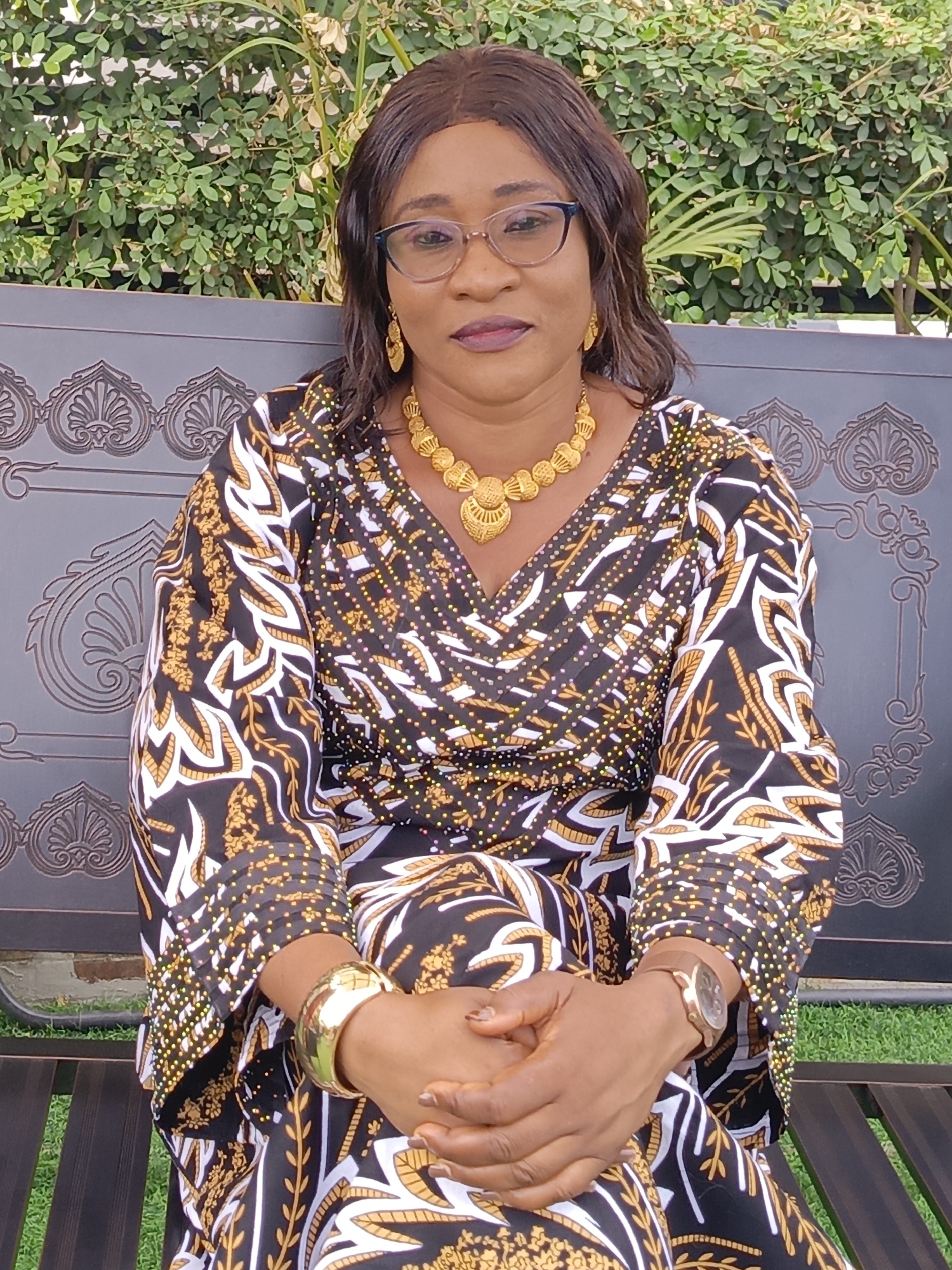 A middle-aged woman is seated outdoors on a decorative dark metal garden bench, set against a backdrop of lush green foliage. She has dark hair, wears dark-framed glasses, and is looking towards the viewer with a calm expression. Her lips are adorned with dark lipstick.
She is wearing a long-sleeved garment, likely a dress or tunic, featuring a striking pattern of gold and white abstract shapes, some resembling leaves, against a dark background. The sleeves are embellished with small, shimmering gold dots. Around her neck, she wears a prominent, ornate gold-colored necklace with multiple dangling elements, complemented by matching gold drop earrings. On her left wrist, she has a wide, polished gold cuff bracelet, and on her right wrist, a watch with a brown strap and a gold-toned casing. Her hands are clasped together in front of her.
The setting appears to be a garden or patio area in Akwanga, Nigeria, bathed in natural daylight, suggesting pleasant weather. There is no visible text in the image.