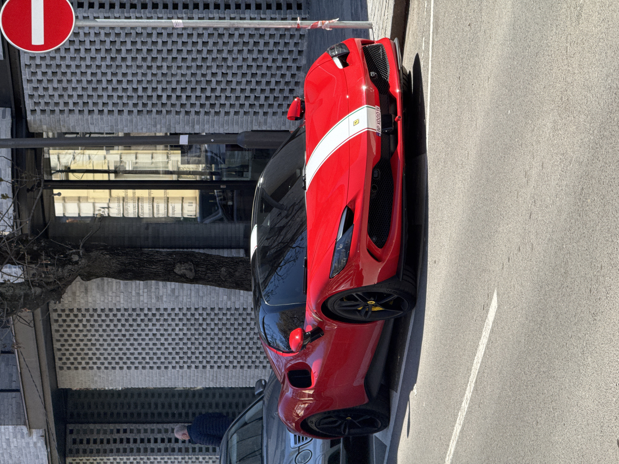 The image shows a red Ferrari SF90 Stradale parked on a city street next to a building with a distinctive grey brick facade. A "No Entry" traffic sign is visible on the upper left. Part of a tree is visible behind the traffic sign and brick building. A person wearing a dark blue sweater and khaki pants can be seen behind the car. The location is identified as Sofia, Bulgaria, based on the license plate "BP X000 TK" on the vehicle.