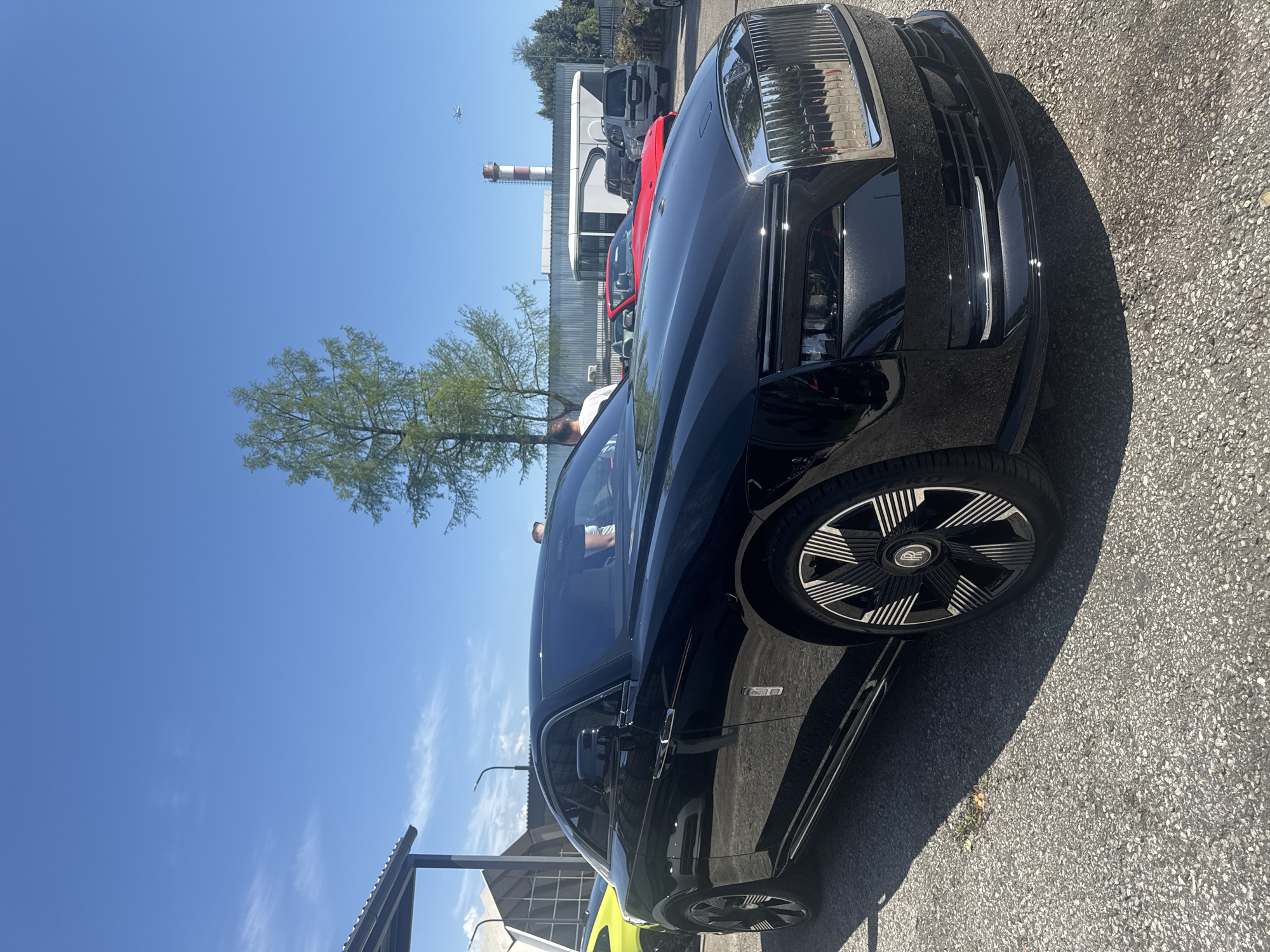 The image shows a black car, possibly a Rolls Royce, parked on an asphalt surface. In the background, there are other cars, a tree, and a building with a tall chimney or tower. A person is visible inside the car. A closer inspection shows the wheel and tires of the vehicle. The location is possibly in or near Sochi, Russia, based on the urban industrial background and the car's likely association with affluent areas, though this is not definitively confirmed within the image itself.