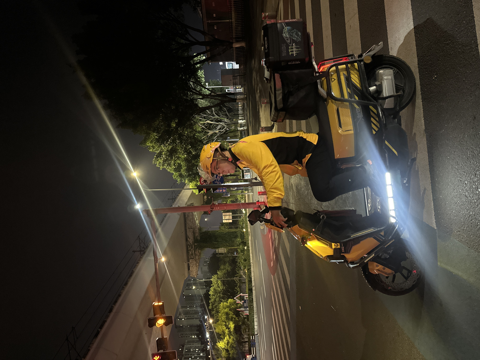 A person wearing a yellow and black jacket and a yellow helmet is standing next to a yellow electric scooter at night. The scooter has a large black delivery box on the back with logos. The person appears to be looking down at the scooter. The scooter is on a pedestrian crossing with black and white stripes. In the background, there are trees, buildings, and streetlights. There is also an elevated road or bridge visible. The scene suggests the person is a delivery driver on a break or waiting at a traffic light. The overall lighting indicates it is nighttime, and the atmosphere is urban. Visible text on the delivery box includes "XDX" and "MONSTER ENERGY". The image is taken in Chongqing High-tech Zone, China.