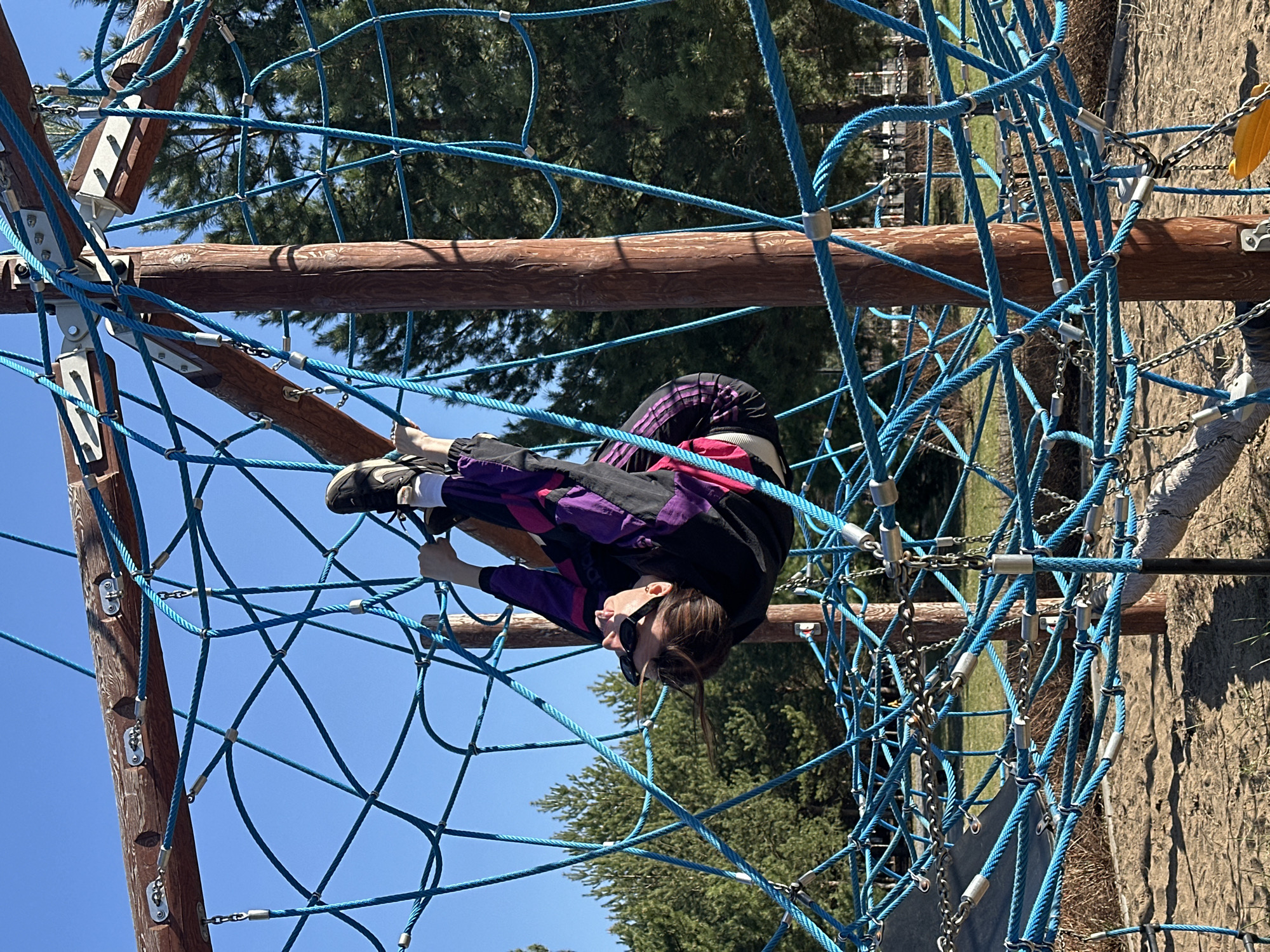 A person is positioned upside down on a blue rope climbing structure. The person is wearing a black jacket with purple and pink accents and black shoes. The climbing structure is constructed from wooden poles and blue ropes arranged in a net pattern. In the background, there are trees and a blue sky. The location is Русский Вожой, Russia.