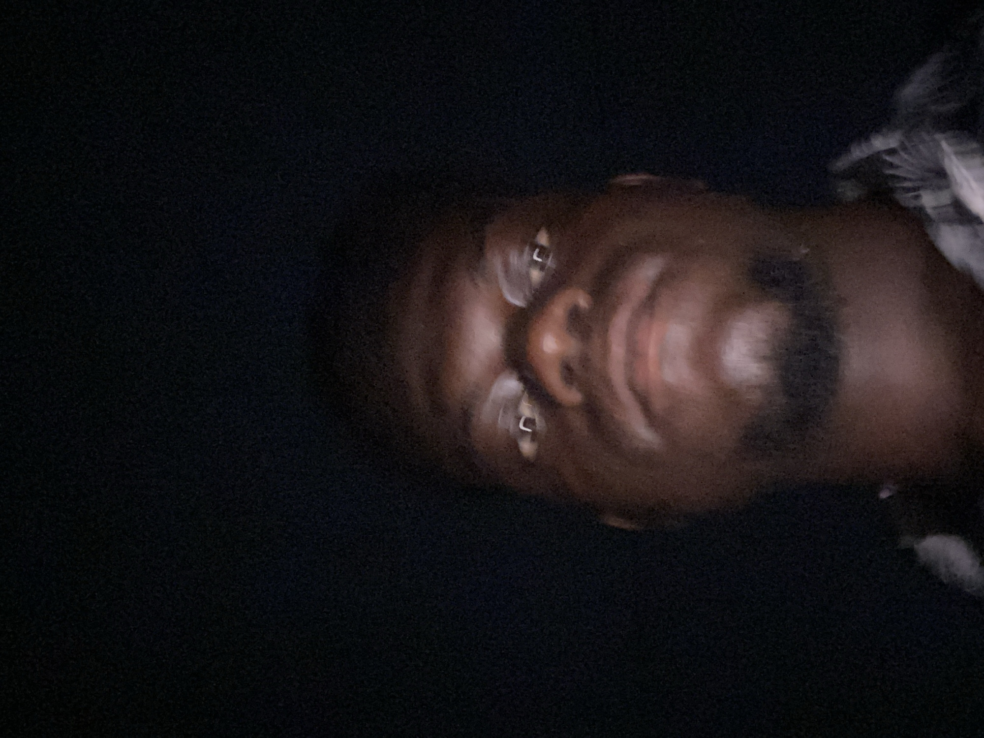 A close-up, sideways photograph captures a person's face against a dark, featureless background, suggesting nighttime or very low-light conditions. The subject is a person with dark skin, appearing to be male, with short hair and a beard or stubble. Their face is partially illuminated, highlighting the forehead, nose, and lips, with distinct reflections visible in both eyes. The person is looking directly at the camera with a slight smile or smirk, conveying a calm and direct gaze. A small section of a patterned or plaid shirt is visible over the person's right shoulder/upper arm on the right side of the frame. The image has a grainy quality, characteristic of photography in dim lighting. No specific location cues or text are visible.