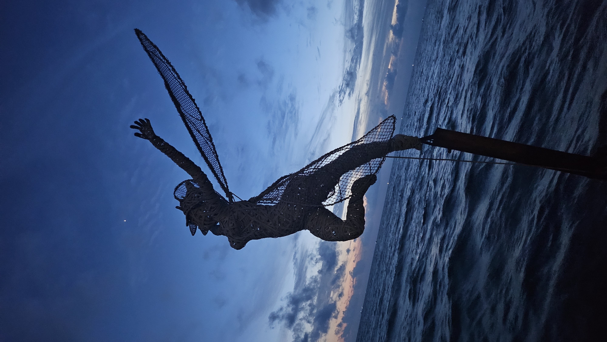 The image depicts a silhouette of a human-like sculpture made of woven material, possibly bamboo or rattan, with sections covered in netting. The sculpture is in a dynamic pose, with one arm outstretched and a net extending from its other side as if it were a wing or sail. It is mounted on a dark, weathered pole, which is partially submerged in choppy, dark blue water. The background is a twilight sky with hues of deep blue, with some wispy clouds near the horizon. A small, bright point, possibly a star or the moon, is visible in the upper part of the sky. The scene suggests an outdoor location, likely near the coast, given the water and sky. The location is identified as the city of Phú Quốc, Vietnam.