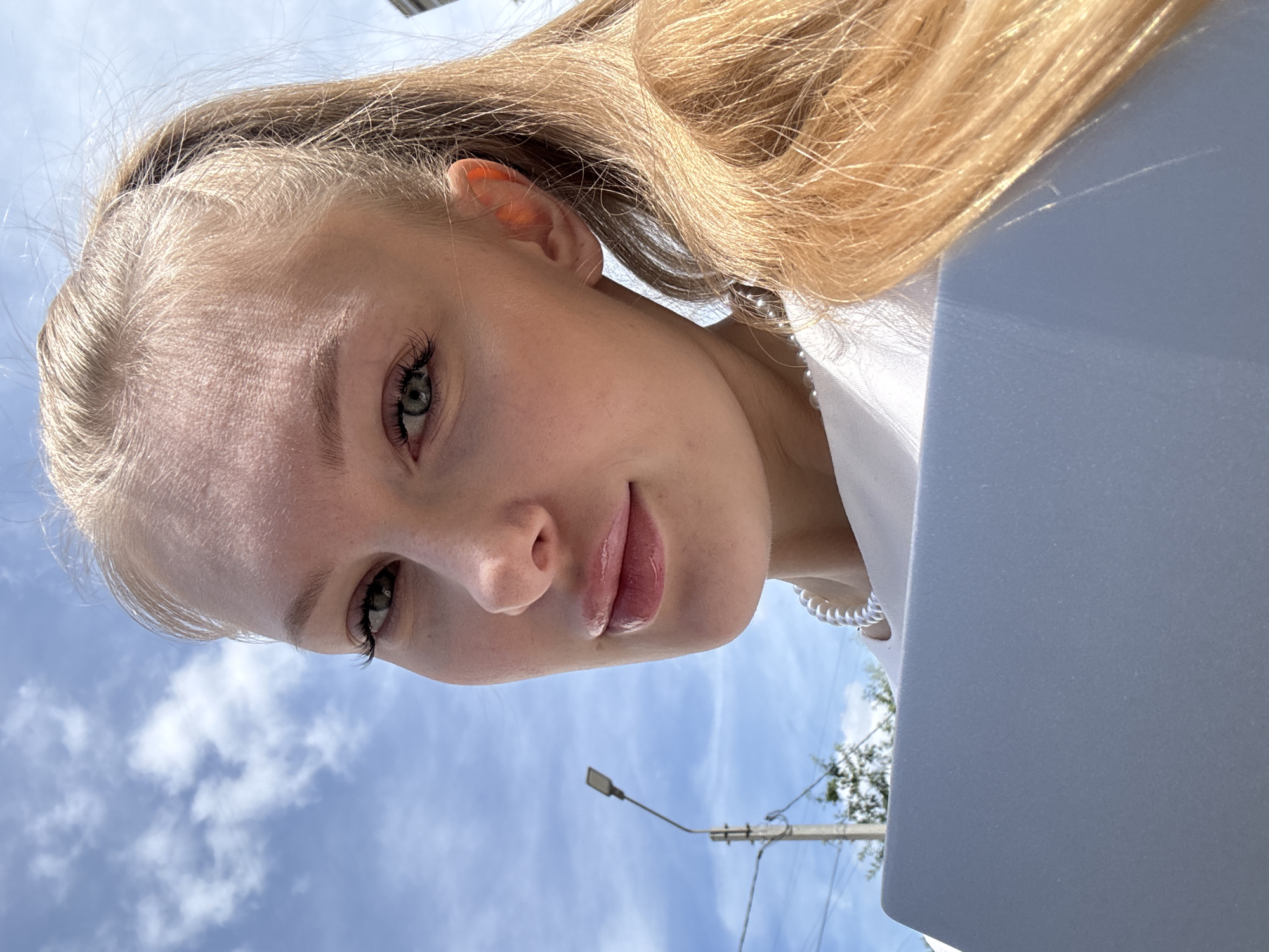 The image presents an upward-angled close-up portrait of a young woman with light blonde hair, fair skin, and green eyes. Her head is slightly tilted, and she gazes directly into the camera with a neutral, perhaps serene, expression, a hint of a soft smile on her naturally glossy, pink lips. Her eyelashes are long and dark.
She is wearing a white garment, possibly a top or dress, and a white, pearl-like necklace is visible on her right side. Her blonde hair frames her face and flows towards the right.
The background is dominated by a bright, partly cloudy blue sky. On the lower left, a telephone pole with a street lamp and some distant green foliage are visible, suggesting an outdoor, urban or suburban setting. On the right side of the frame, a large, light grey or white object, possibly a laptop or tablet, is partially visible, reflecting some light.
The scene is set outdoors during the daytime, under bright natural light, indicating clear weather. The low camera angle gives the impression of a selfie or a photo taken from below. The overall mood is bright and fresh, set against the backdrop of Perm, Russia. No text is visible in the image.
