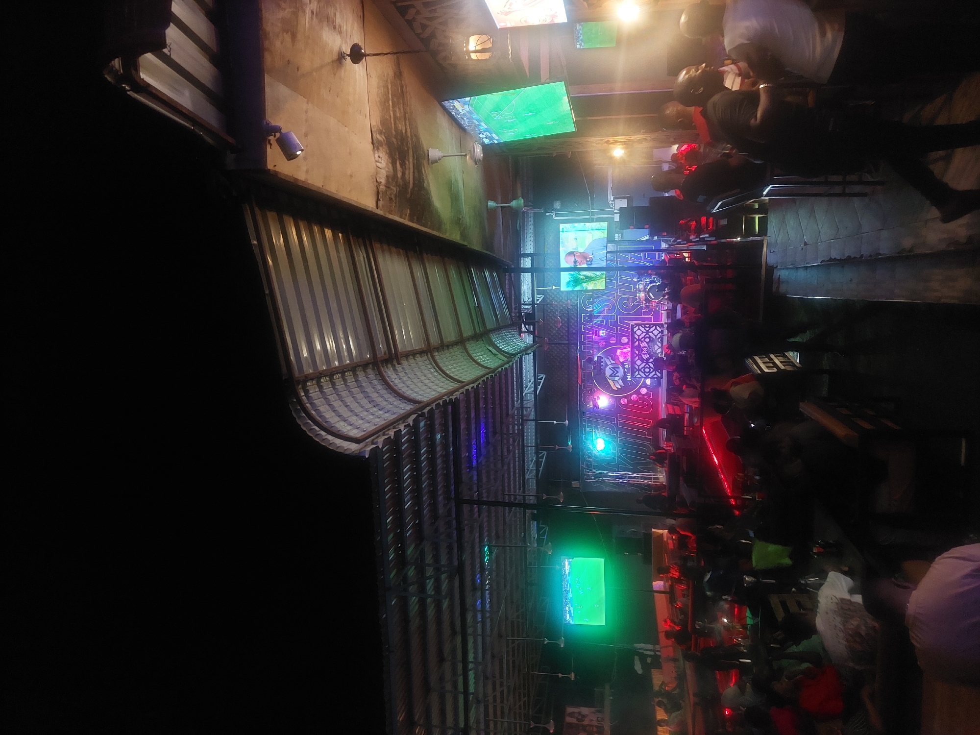 The image depicts an outdoor bar or restaurant in Dar es Salaam, Tanzania, at night. The scene is crowded with people, some standing and others seated at tables. Several televisions are mounted on the walls, displaying sports games. Lighting is provided by overhead fixtures and colorful neon signs, creating a lively atmosphere. The building is a mix of open-air and covered spaces with visible building materials. There are people interacting and watching the sports game.