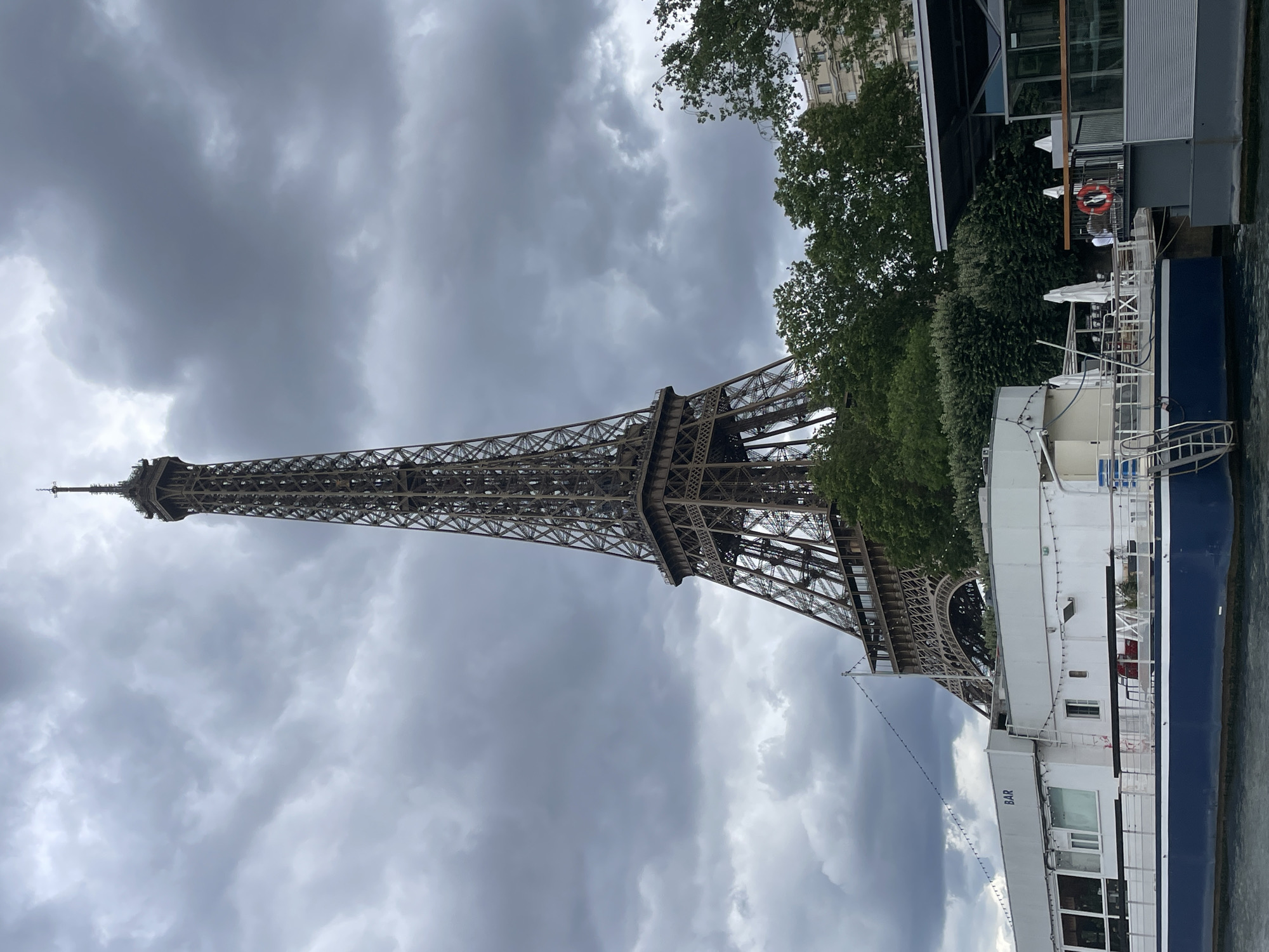 The image shows the Eiffel Tower in Paris, France, under a cloudy sky. In the foreground, a blue and white boat with a "BAR" sign is docked on the Seine River. The boat has a white upper deck with windows, ladders, and tables. To the right of the boat, a modern structure with large windows and a railing is visible, with a red life preserver hanging on the railing. Trees and buildings are in the background, partially obscured by the Eiffel Tower and the trees.