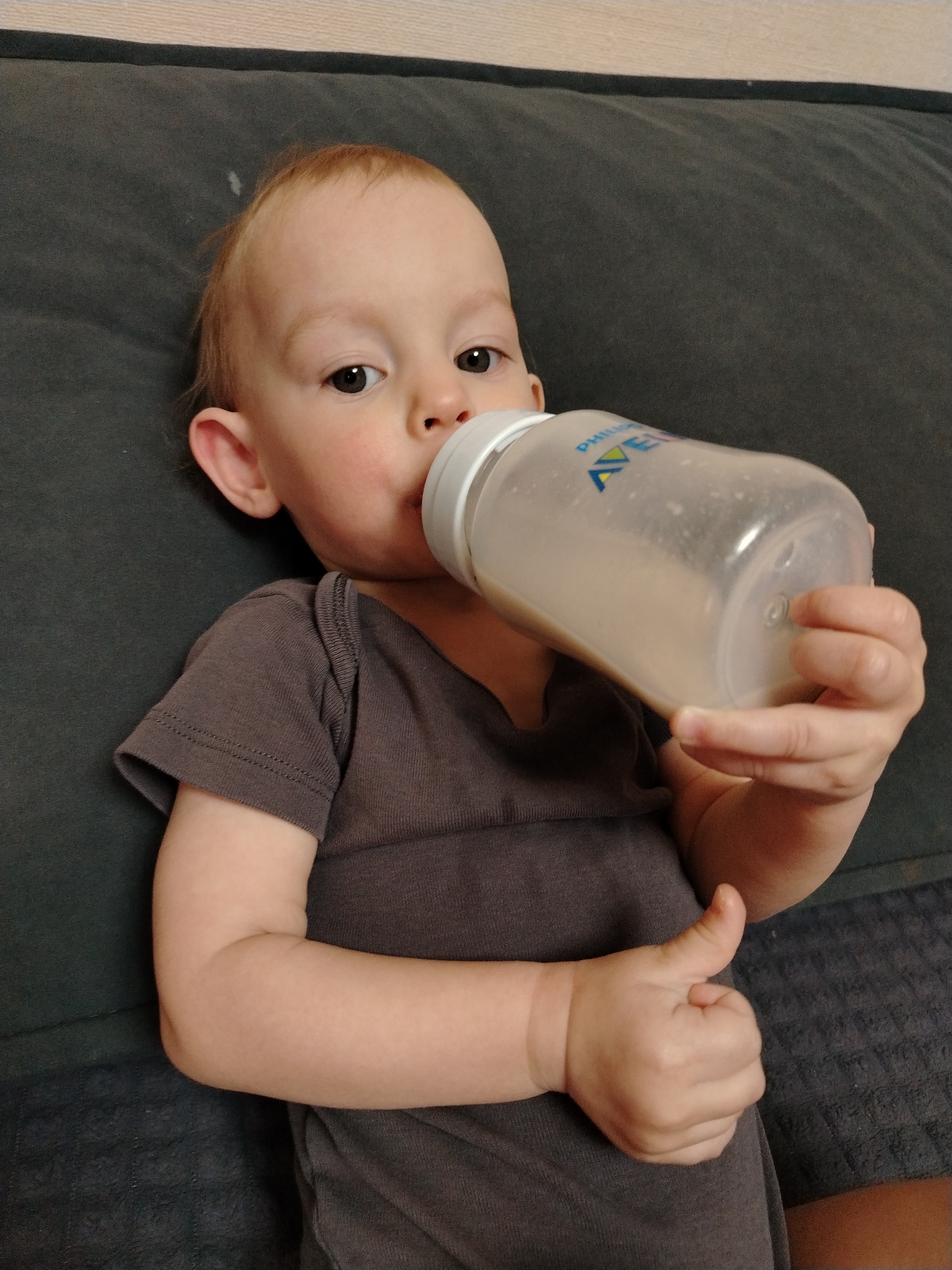 A baby lies on a dark gray couch or surface, wearing a gray shirt and drinking from a Philips Avent bottle. The bottle contains a light-colored liquid, possibly milk or formula. The baby is holding the bottle with both hands and appears to be looking at the viewer. No location of Chaikovsky, Russia is visible in the image.