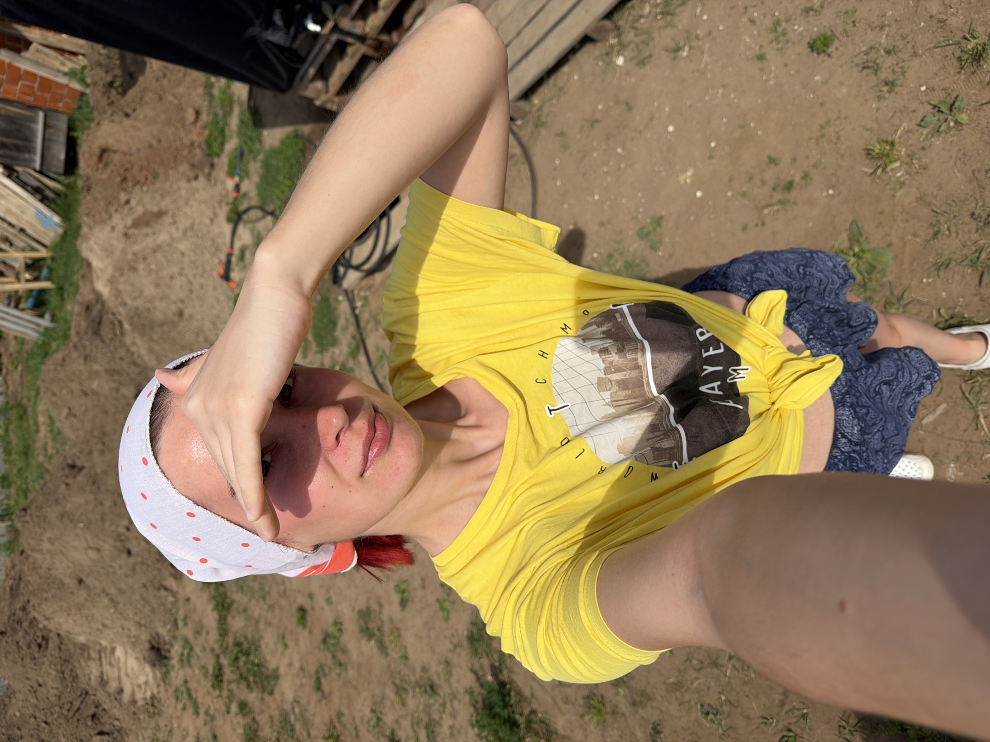 A woman wearing a yellow T-shirt with a building illustration, a blue patterned skirt, and white shoes is lying on the ground. She is wearing a white headscarf with red polka dots. She is holding her hand to her forehead. The ground appears to be a mix of dirt and grass. In the background, there is a brick building, some wooden structures, and various objects on the ground. The image was taken at an unspecified location in Chaikovsky, Russia.