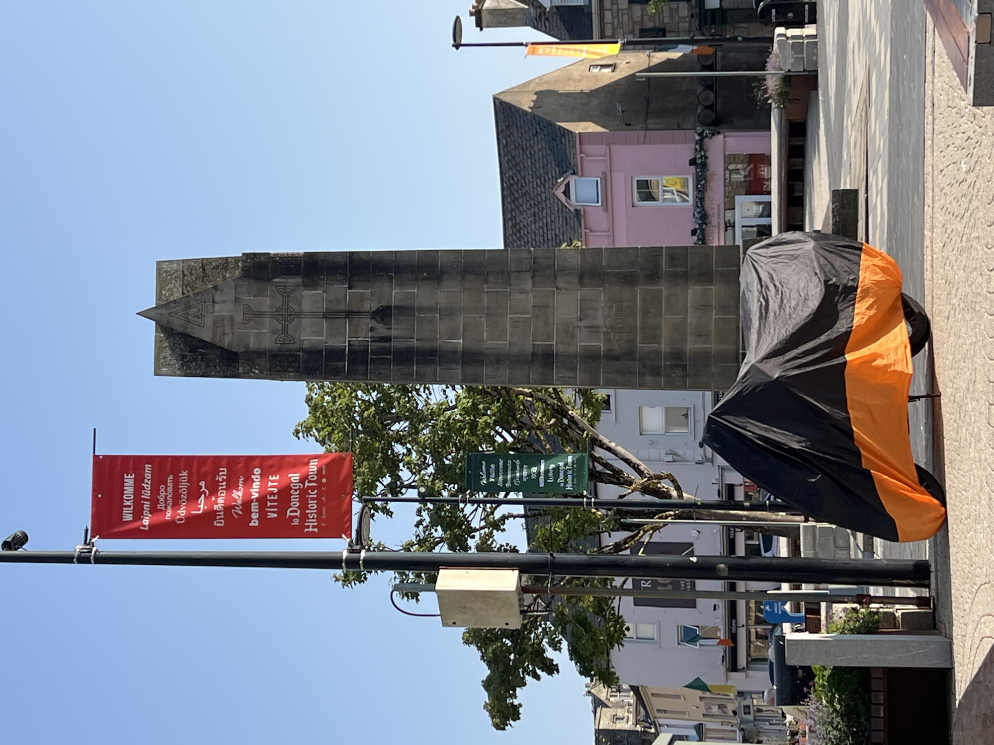 The image shows a street scene in Donegal Town, Ireland. In the foreground, a motorcycle is covered with a black and orange tarp. Behind the motorcycle, a large stone obelisk stands tall, adorned with a Celtic cross design. Red and green banners hang from lampposts, displaying the town's name in multiple languages. Buildings with pastel-colored facades line the street, some featuring businesses with signage. The sky is clear and blue. The overall scene depicts a typical day in a historic Irish town.