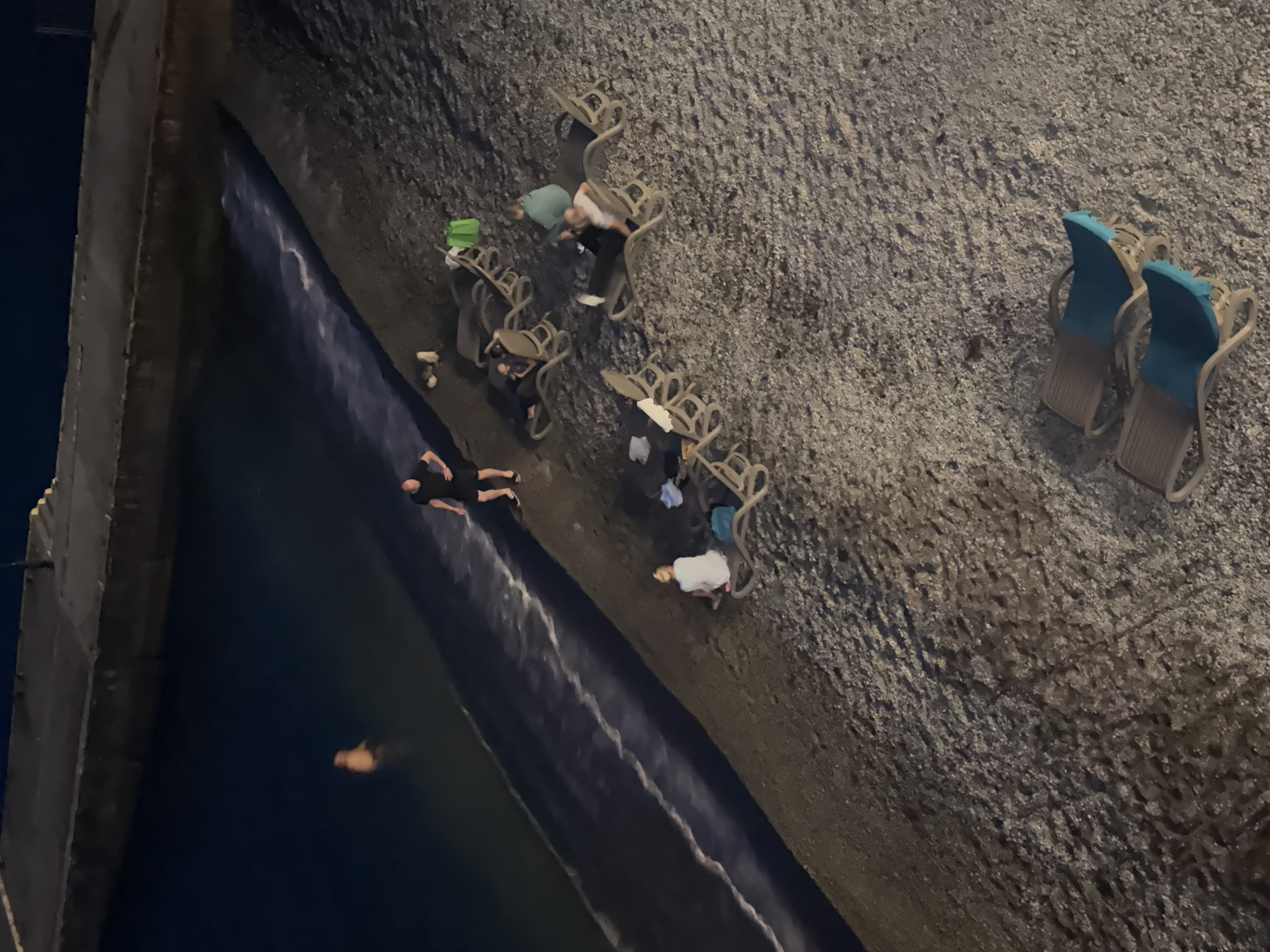 In the city of Yalta, Ukraine, a beach scene unfolds at night. The water's edge meets a pebbled shore where several lounge chairs are set up. A person is partially submerged in the dark water near a concrete embankment. On the beach, a man stands by the water, while further inland, several individuals are seated or lounging on chairs. A green bag is visible near some of the chairs. In the foreground, two lounge chairs with blue cushions are unoccupied. The lighting suggests it is nighttime, with artificial light sources illuminating parts of the scene.
