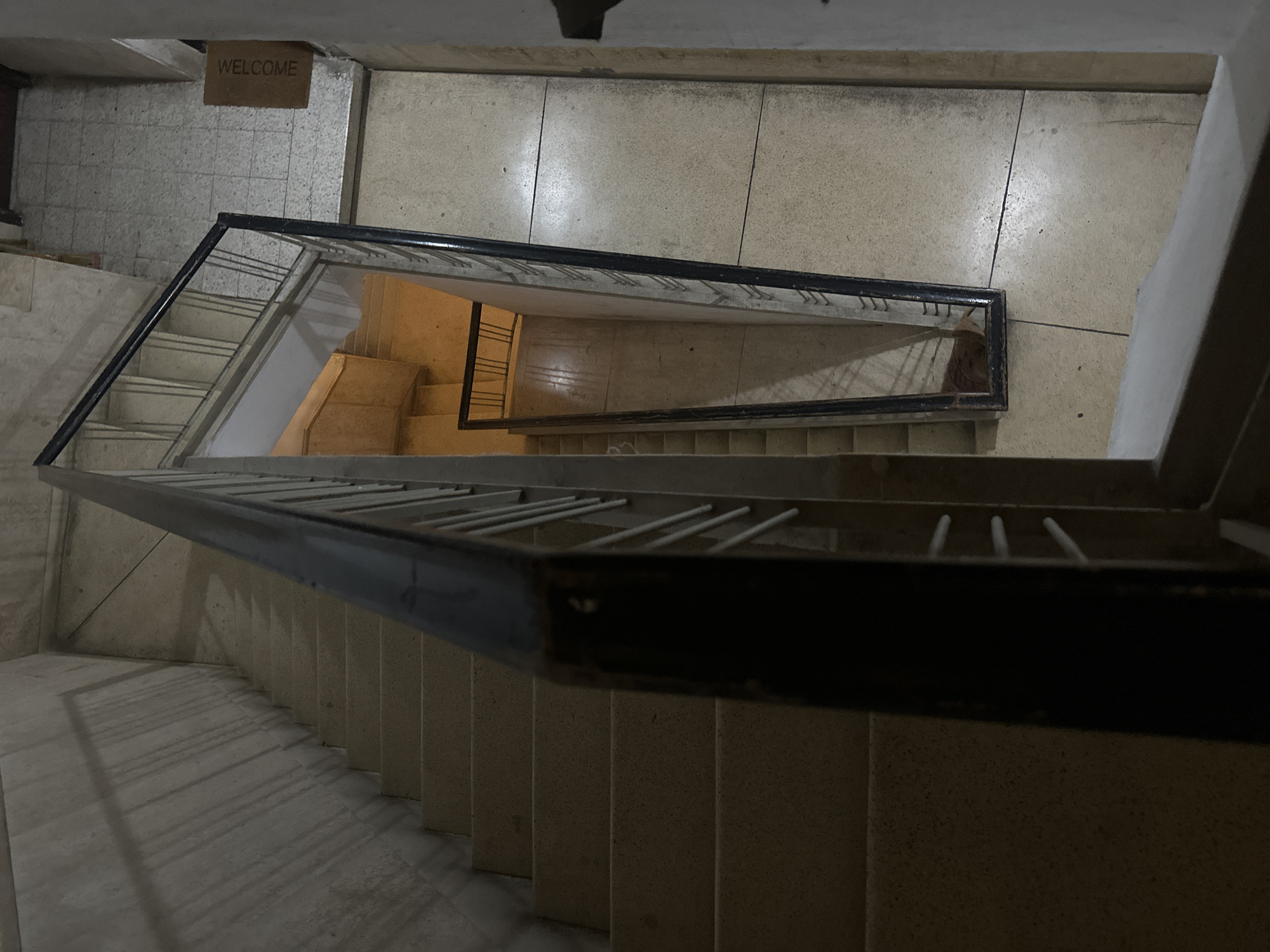 An interior shot captures a view looking down a staircase in an apartment building in Tel-Aviv, Israel. The staircase is made of concrete with a dark railing. On the right side of the image, a "WELCOME" doormat is visible on the tiled floor. The lighting suggests it is either early morning or late evening.