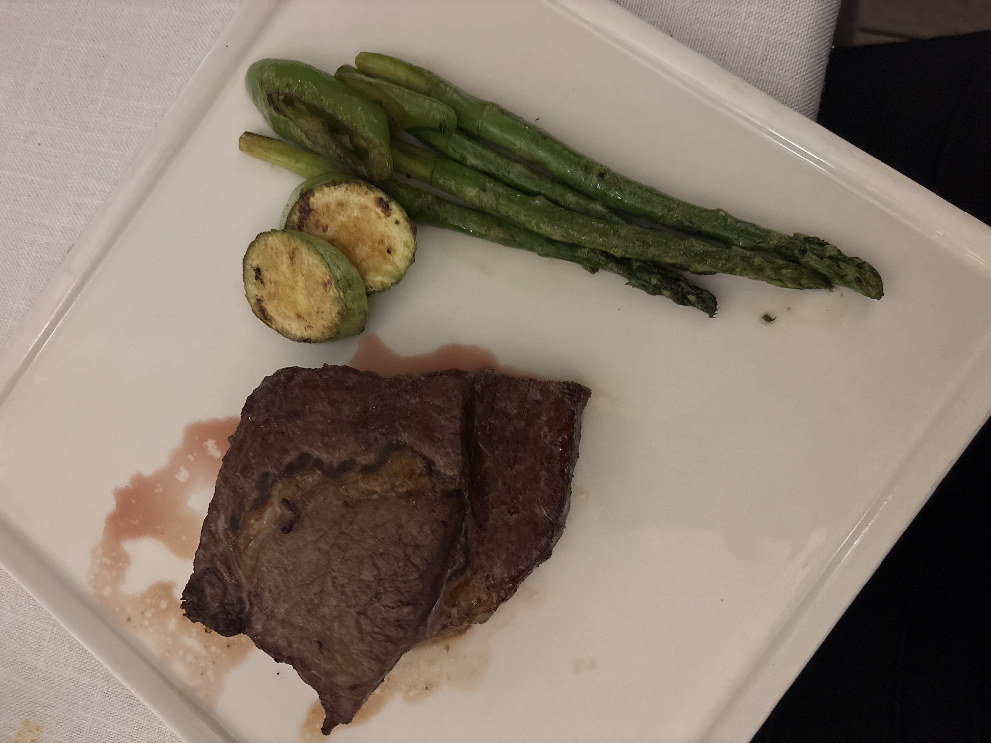 A grilled steak is presented on a white rectangular plate alongside grilled zucchini slices and asparagus. The steak is a deep brown with some reddish-pink juices on the plate. The zucchini slices are light brown with visible grill marks, and the asparagus spears are a vibrant green, also showing signs of grilling. The plate is set on a white tablecloth. The scene is likely from a restaurant in Varna, Bulgaria, given the context of the request.