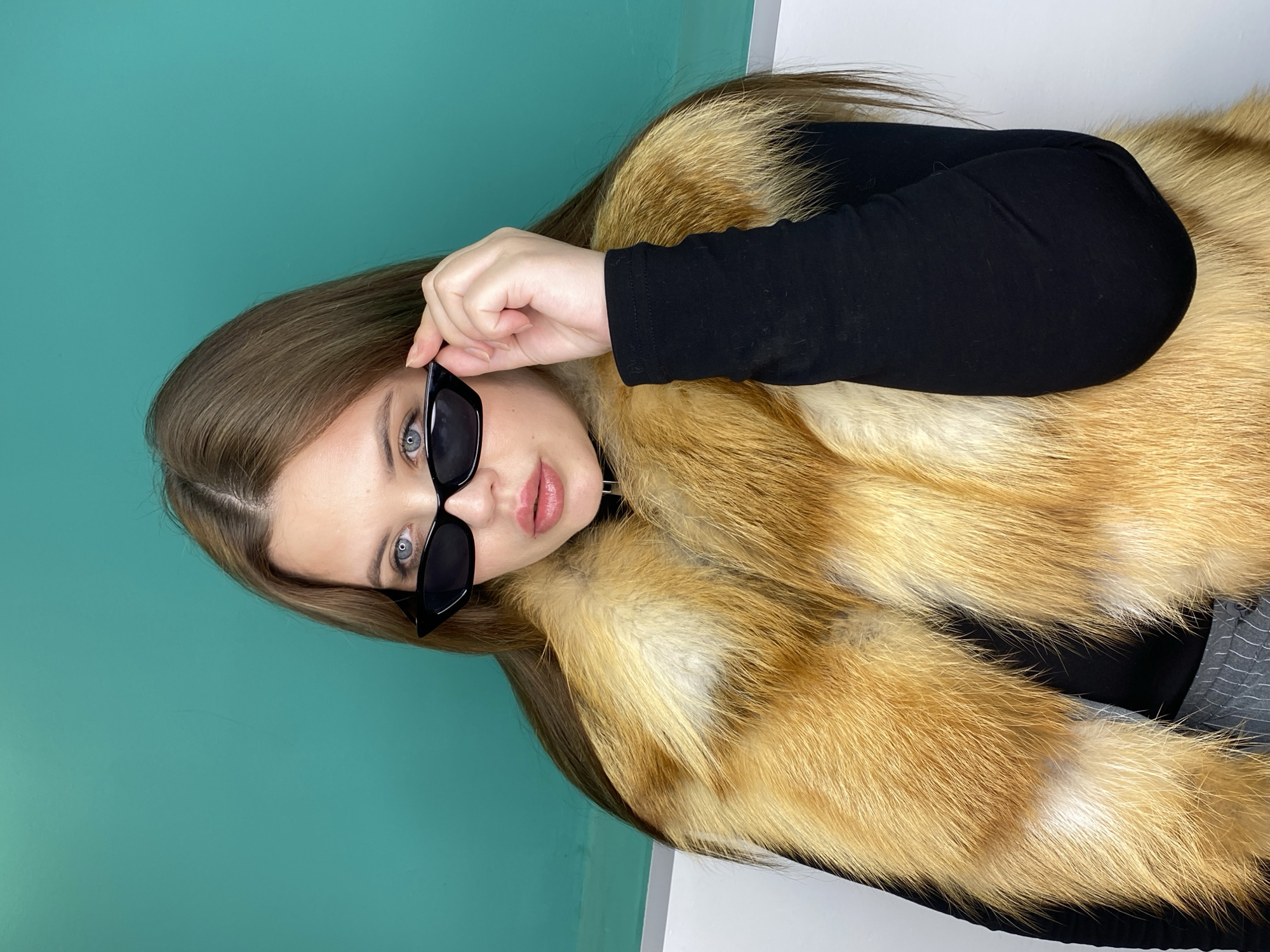 A young woman with light brown hair and blue eyes is posing indoors. She is wearing a black long-sleeved shirt and a large, golden-brown fur coat. She is also wearing black sunglasses and is holding the frame of the sunglasses with her right hand, pulling them slightly down her face. The background is a two-toned wall, with the top half being a light teal color and the bottom half a bright white. The location and date are unknown.