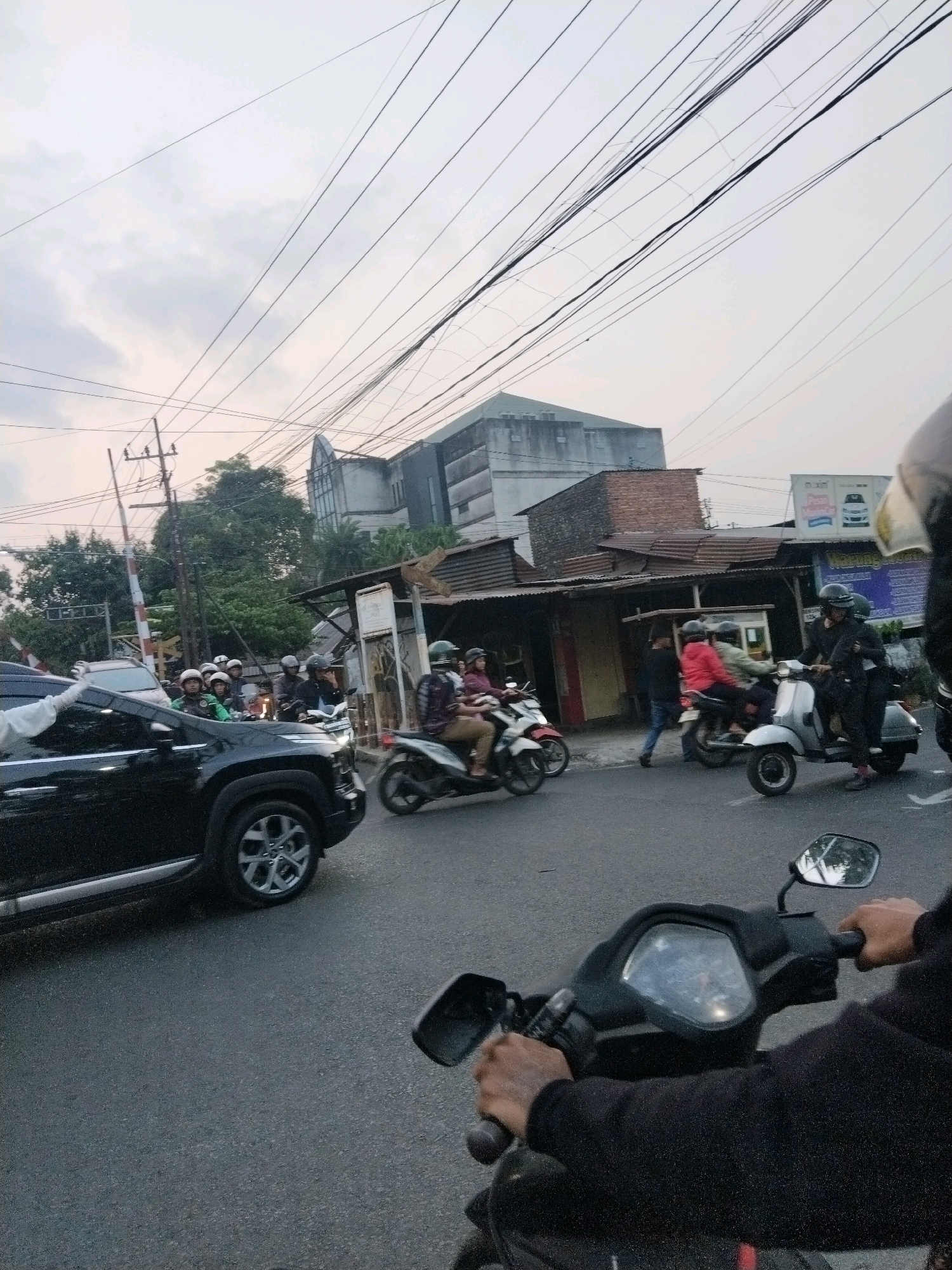 The image depicts a busy street scene in Malang, Indonesia, likely near a railway crossing given the presence of a barrier. A black SUV is stopped in the foreground, on the left side of the frame, with its wheel visible. Several motorcycles are in motion and stopped on the road. One motorcycle in the foreground, from the perspective of its rider, shows the handlebars and a side mirror. On the right, a person in black is riding a scooter, and behind them, another scooter with two riders is visible. Further back, a group of people on motorcycles are waiting, some wearing helmets. The background shows a building with a distinctive architectural style, likely a public building or place of worship, and other smaller structures. Overhead, a complex network of power lines crisscrosses the sky, which is overcast. The scene suggests moderate traffic flow in an urban environment.