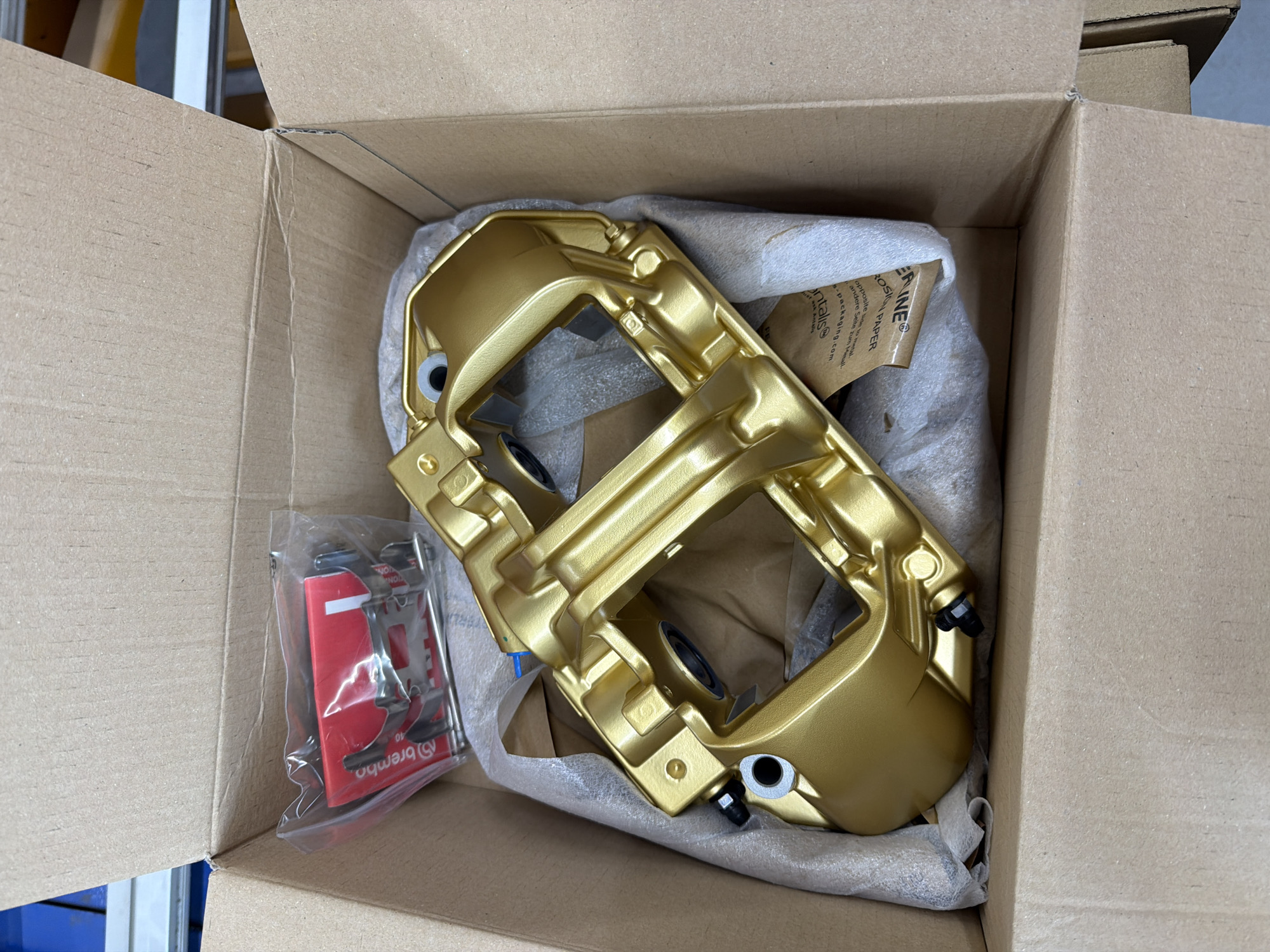 The image shows a golden brake caliper in a cardboard box. Next to the caliper is a package with red and silver Brembo brake pads. The caliper is surrounded by crinkled, waxy packaging paper. A piece of packaging paper is visible, with the words "ERINE" and "ON PAPER" printed on it, along with text in both English and German regarding the use of the packaging material. The box appears to be new and clean.