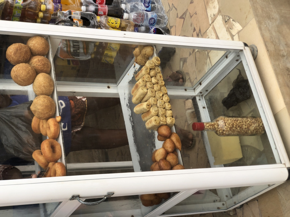 The image depicts a mobile glass display cabinet positioned on a paved surface in Egan, Nigeria. The cabinet, constructed with a white frame and transparent glass panels, features three shelves filled with food items.
On the top shelf, approximately eight golden-brown ring-shaped doughnuts are arranged on the left, while five spherical, golden-brown fried dough balls are stacked on the right. Below this, the middle shelf displays a pile of irregular, golden-brown fried items, possibly fritters, on the left. To their right, a pyramid stack of approximately 30-40 light golden-brown rolled items, resembling spring rolls, occupies a significant portion of the shelf. A smaller cluster of additional fried items is visible further right. The bottom shelf contains a large, pale yellow rectangular block on the left, a clear glass bottle with a red cap filled with groundnuts (peanuts) in the center, and a dark, crumpled object, possibly a plastic bag, on the right.
Reflections within the cabinet glass show the lower torso and legs of at least two individuals. One person, wearing dark shorts with a patterned hem, is visible behind the top and middle shelves, with their feet appearing on the paved ground. Another set of feet, clad in sandals, is reflected near the bottom shelf. A hand is partially visible in the top left corner, seemingly interacting with items inside the cabinet.
To the right of the display cabinet, an adjacent stall or display area is visible, featuring various bottled beverages including water, Coca-Cola, Fanta, and Maltina, some packaged in clear plastic bags. The ground beneath the cabinet and extending to the right is composed of irregularly shaped tan and brown paving stones.