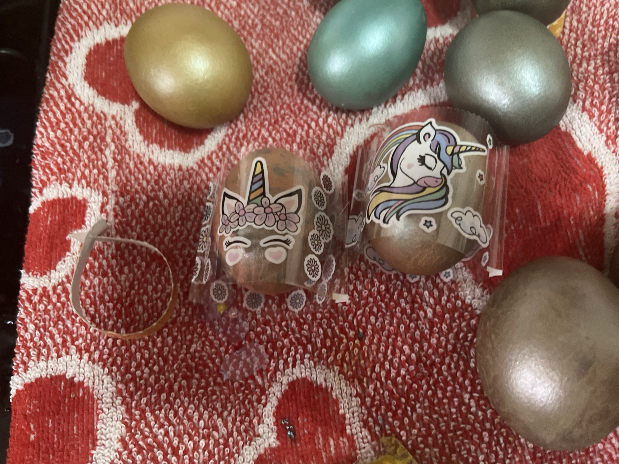 The image shows several decorated Easter eggs resting on a red and white patterned towel. Some of the eggs are plain, painted in gold, teal, and silver. Two eggs are encased in clear plastic shrink wrap with unicorn designs printed on them. One shrink wrap is partially removed. The towel is textured with a looped pattern. The image appears to be taken indoors. There is no evidence to suggest the location of Oryol-Izumrud, Russia, or any specific location.
