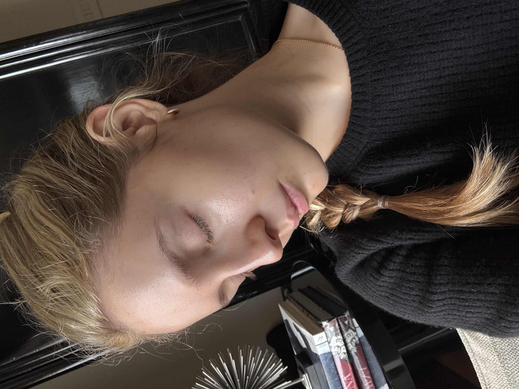 A young woman with fair skin and blonde hair is lying down, likely asleep, on a black surface, possibly a table. Her hair is styled in a braid that is partially visible next to her black sweater. She has a delicate chain necklace and a small earring. In the background, there are books and a metallic object resembling a sea urchin. The location is unclear, and there is no definitive evidence indicating it is Mexico City.