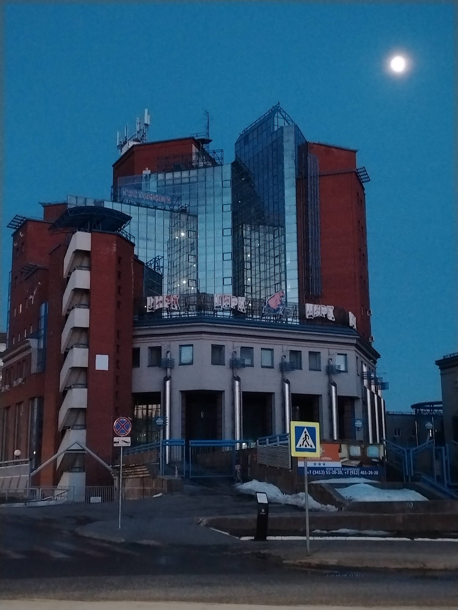 The image is a night shot of the "Arena" Hotel in Izhevsk, Russia, under a moonlit sky. The multi-story building is a mix of red brick and modern glass construction, with the word "GOSTINIMSA" (hotel) visible atop a glass section. Below, the letters "LIRK" are visible in Russian.
Street signs, including a pedestrian crossing sign, are prominently displayed in the foreground. Stairs with blue railings lead up to the building's entrance. Patches of snow are visible on the steps. The street in the foreground appears wet, reflecting the ambient light. Telephone numbers are posted on the front of the stairs leading to the building.