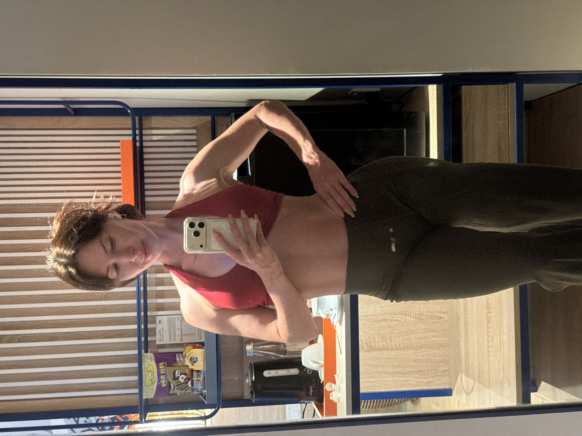 In a hotel room, a woman with brown hair pulled back is taking a mirror selfie. She is wearing a red halter top and dark green leggings. The room has striped wallpaper and wooden shelves. On one shelf, there is a black electric kettle, a bottle of water, and two cups. The woman is holding a smartphone in her right hand, and her left hand is on her hip. The image does not provide any specific details about the location in Dong Ninh Hoa Ward, Vietnam.