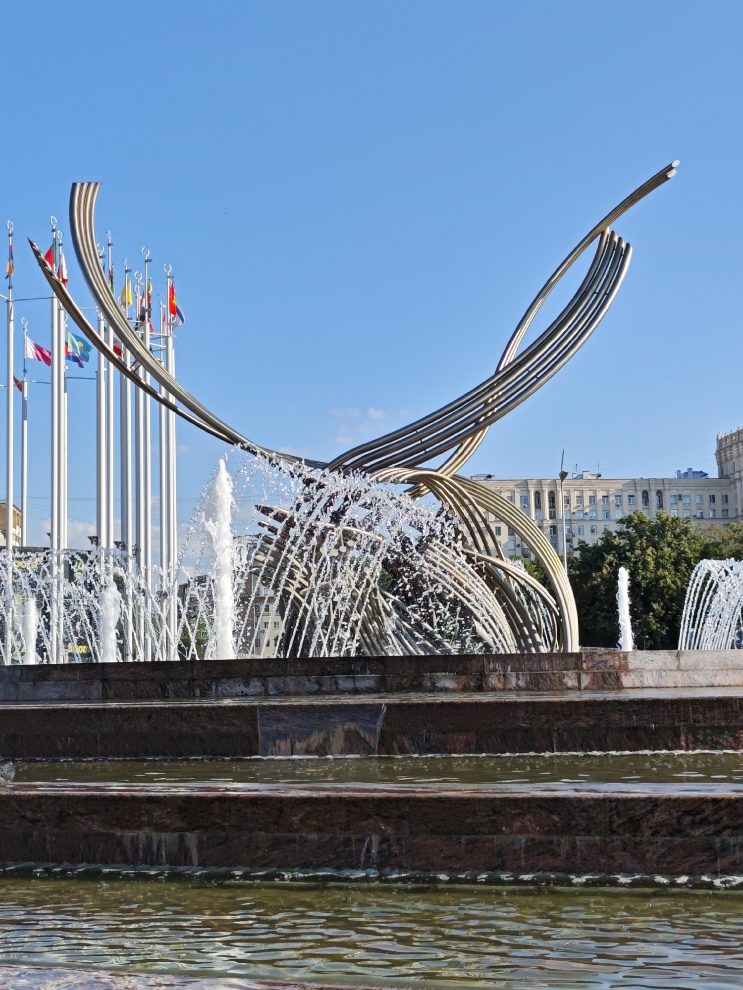 A wide shot captures a modern fountain sculpture with a clear blue sky in the background. The sculpture, made of polished metal, features an abstract, flowing design with multiple curved elements that create a sense of movement. Water sprays upwards from various points within and around the sculpture, creating a dynamic display of water jets and mist.
In the middle ground, rows of tall flagpoles stand in a semi-circle, each flying a national flag. These flags represent various countries, suggesting the location is a place of international significance. Behind the flagpoles, a large, multi-story building with a pale facade and numerous windows is visible. Green trees with full foliage are scattered in front of and around the building, adding a touch of nature to the urban setting.
The foreground consists of a multi-tiered fountain basin made of dark, reddish-brown stone with a polished finish. Water fills the lower levels of the basin, reflecting the light and creating ripples on its surface. The overall scene depicts an urban public space, likely a plaza or park, designed to be visually striking and possibly a landmark. The image's composition, with the sculpture as the central focus and the city architecture in the background, suggests a grand and somewhat monumental setting.
This scene is located at the Monument to the Conquerors of Space in Moscow, Russia. The sculpture represents a rocket ascending into space, and the fountain is a significant part of the monument's design. The flags represent various nations, commemorating the achievements of space exploration.