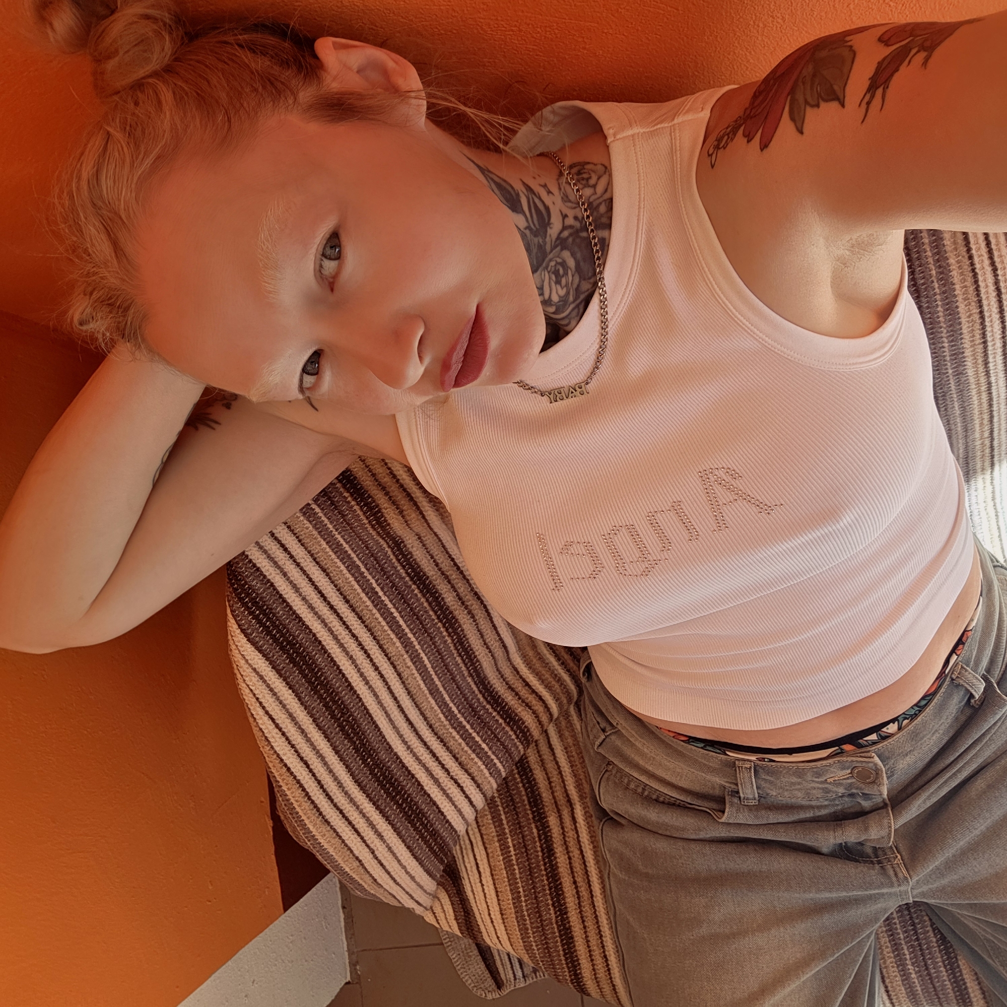 A young woman with blonde hair styled in a bun is lying on a striped blanket over what appears to be a vibrant orange surface. She is wearing a white tank top with the word "Angel" spelled out in rhinestones and a pair of faded denim jeans. Tattoos are visible on her neck, upper arm, and forearm. She is wearing a necklace that says "BABY". Her head rests on her left hand, and she is looking up at the camera. The setting may be indoors. The location provided, Тэльмінскі сельскі Савет, Belarus, is not directly evident from the image itself.