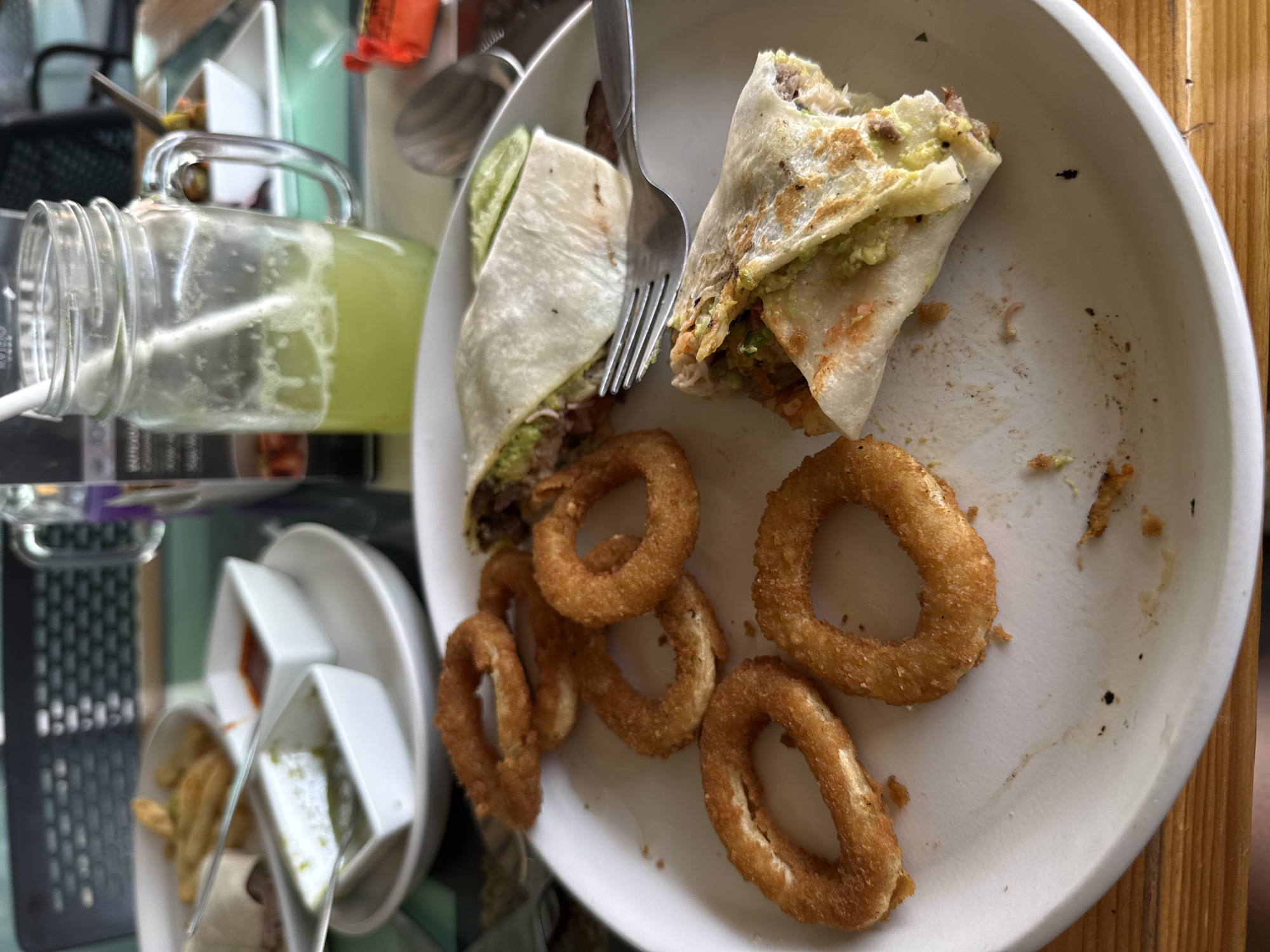 The image captures a partially consumed meal on a white plate, consisting of two halves of what appear to be stuffed tortilla wraps, along with several onion rings. A fork is piercing one of the wrap halves. To the left of the plate, there's a mason jar filled with a yellow-green liquid, likely a beverage. Further to the left, a small dish contains what looks like fries and possibly condiments in small square containers. The table appears to be made of light-colored wood. The scene suggests a restaurant or cafe setting. It's not possible to determine the location in Puebla, Mexico from this image.
