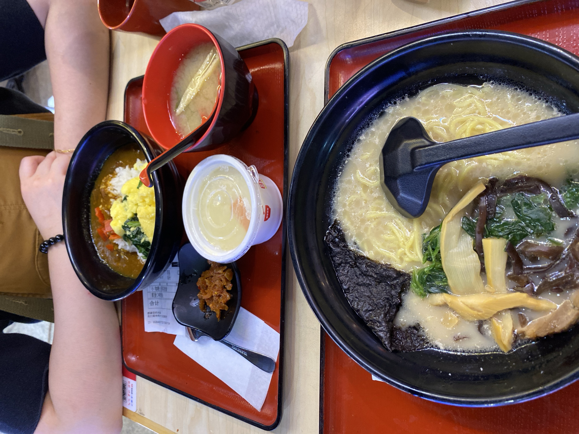 This is a close-up, eye-level shot of two Japanese meals on red trays on a wooden table. The meals are placed side-by-side. The tray in the background contains a bowl of curry rice with scrambled eggs and spinach, a small dish of pickled vegetables, and a bowl of miso soup. A person's arms are visible in the upper part of the frame. The tray in the foreground contains a bowl of creamy broth ramen with noodles, seaweed, mushrooms, and green vegetables. A black ladle rests in the ramen. This scene is at a restaurant located in Qingyang District, China.