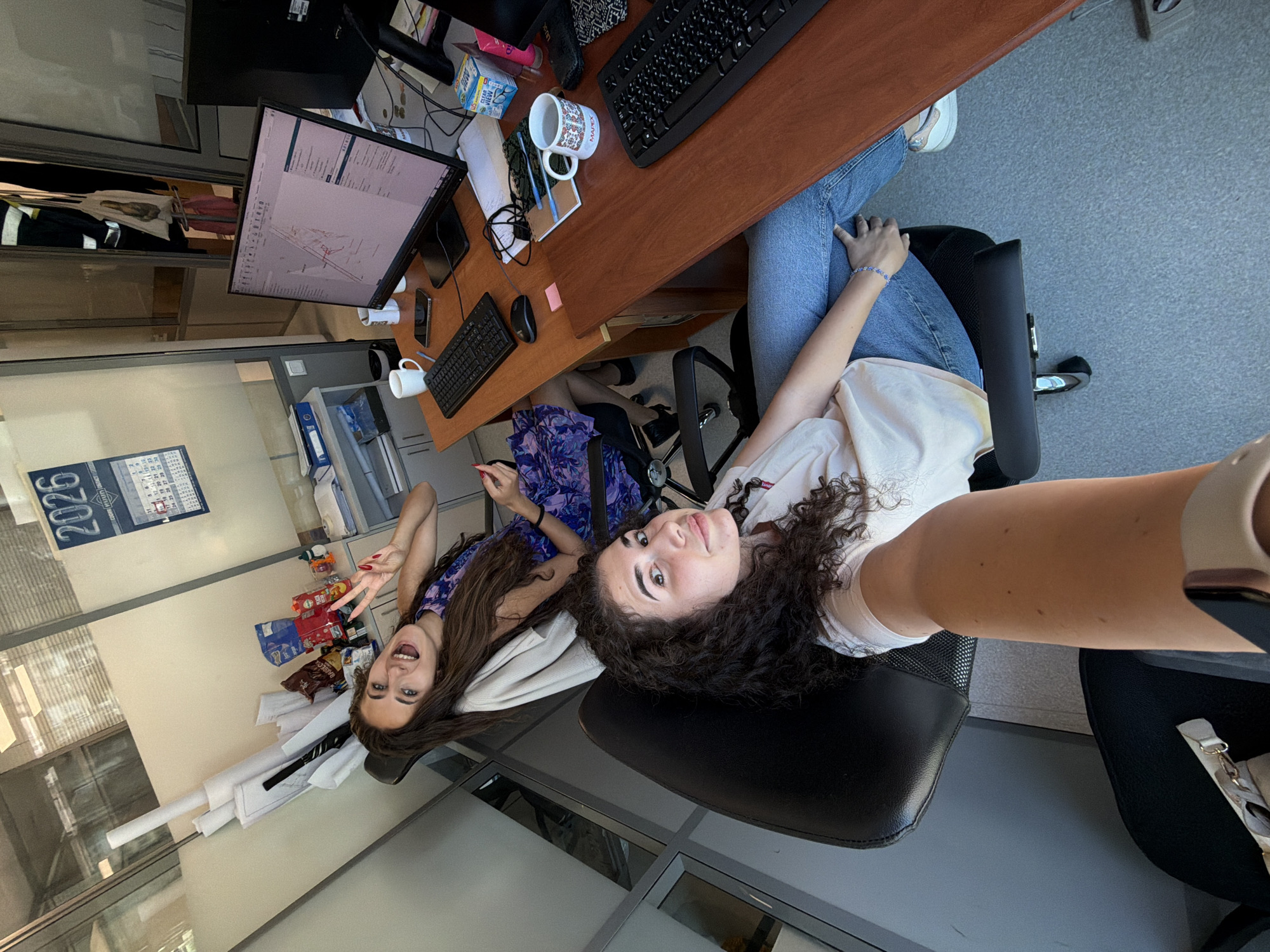Two young women are seated at desks in an office environment. The woman in the foreground is facing the camera, with curly dark hair, wearing a white t-shirt and blue jeans. She is seated in a black office chair. The woman behind her has long, dark hair and is wearing a purple floral dress. She is also seated in a black office chair and is making a peace sign with her hand, looking at the camera and smiling. The office appears to be well-lit and contains typical office furniture and equipment, including computers, keyboards, a monitor displaying a technical drawing, stacks of papers, and office supplies. A calendar for the year 2026 is visible on the wall. The overall impression is one of a casual, friendly workplace. The image does not provide any specific clues to pinpoint its location within the city of Sofia, Bulgaria.