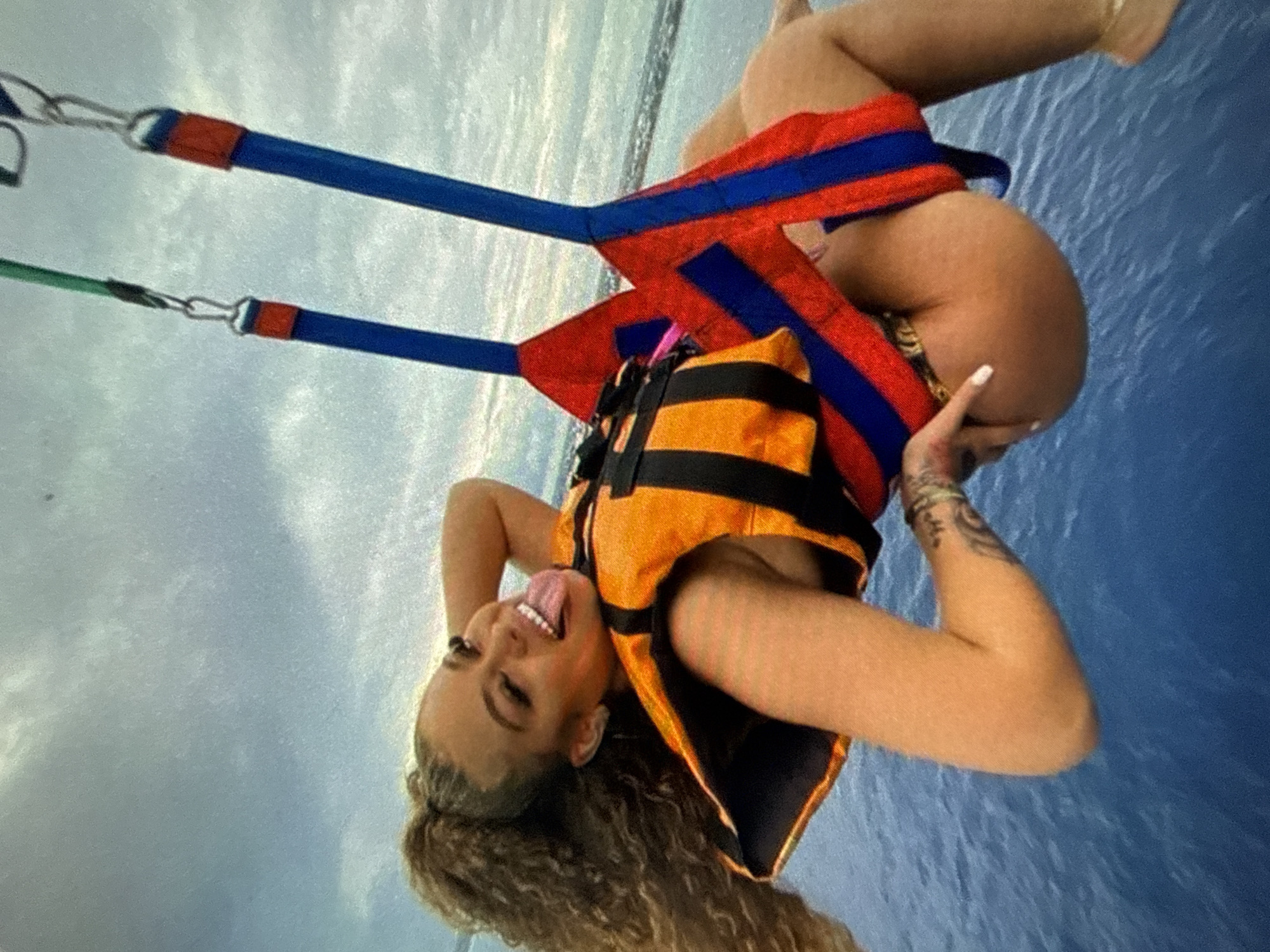 A woman is suspended upside down in a parasailing rig, likely over the ocean, given the water in the background. She is wearing an orange and black life jacket, a red and blue harness, and a bikini or swimwear underneath. The woman is smiling with her tongue sticking out, suggesting a playful or humorous attitude. She has fair skin, curly brown hair, and a visible tattoo on her left arm. The parasailing rig has blue straps with red sections near the metal connectors. The sky is overcast, providing a diffused light to the scene. The city is unknown.