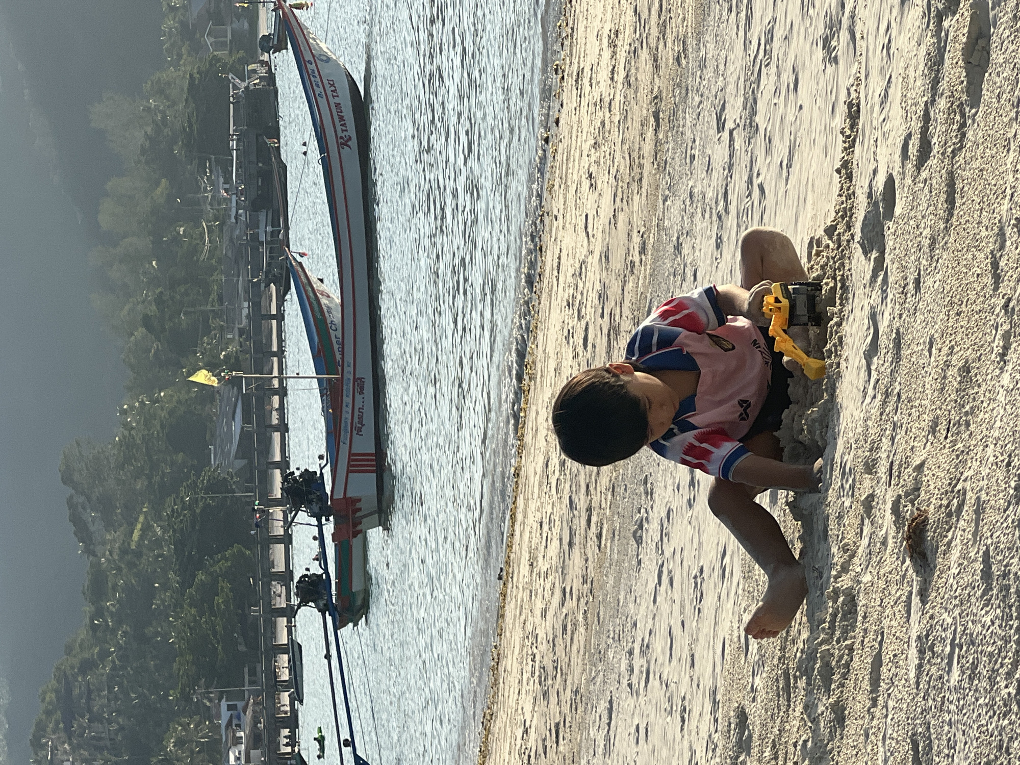 In Ko Pha-ngan District, Thailand, a young boy sits on a sandy beach, playing with a yellow toy excavator. He is wearing a pink t-shirt with red and blue accents. Behind him, the ocean is calm, with several longtail boats moored. The boats are white with red and blue trim, and one is labeled "TAWUN TAXI." In the background, a lush green hillside rises, dotted with buildings and vegetation. The sun appears to be setting, casting a warm glow over the scene. The beach is mostly empty, with only a few distant figures visible near a pier in the far distance. The tide is low, revealing damp sand and small shells.