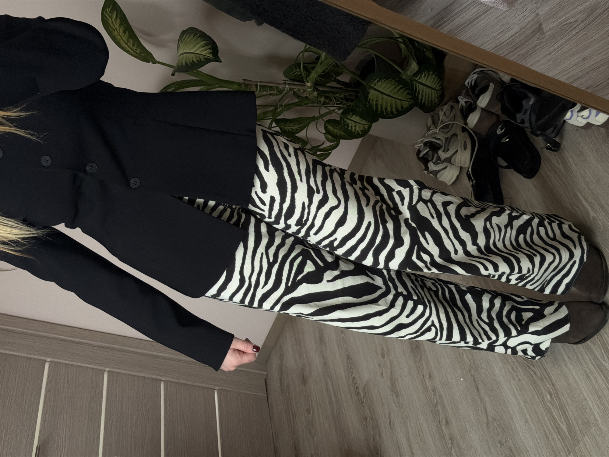 The image shows a person wearing a black blazer and zebra-print pants, standing on a light brown floor. The person is facing forward, with part of their blonde hair visible on the left. A plant with green and white leaves is visible in the upper center of the image. Other objects, including shoes, are visible in the top right corner, underneath what appears to be a table or shelf. The background consists of a wall with wooden-like panels on the lower left. There's no specific indication of the city of Dreiliņi, Latvia in this image, therefore it would be impossible to confirm this fact.