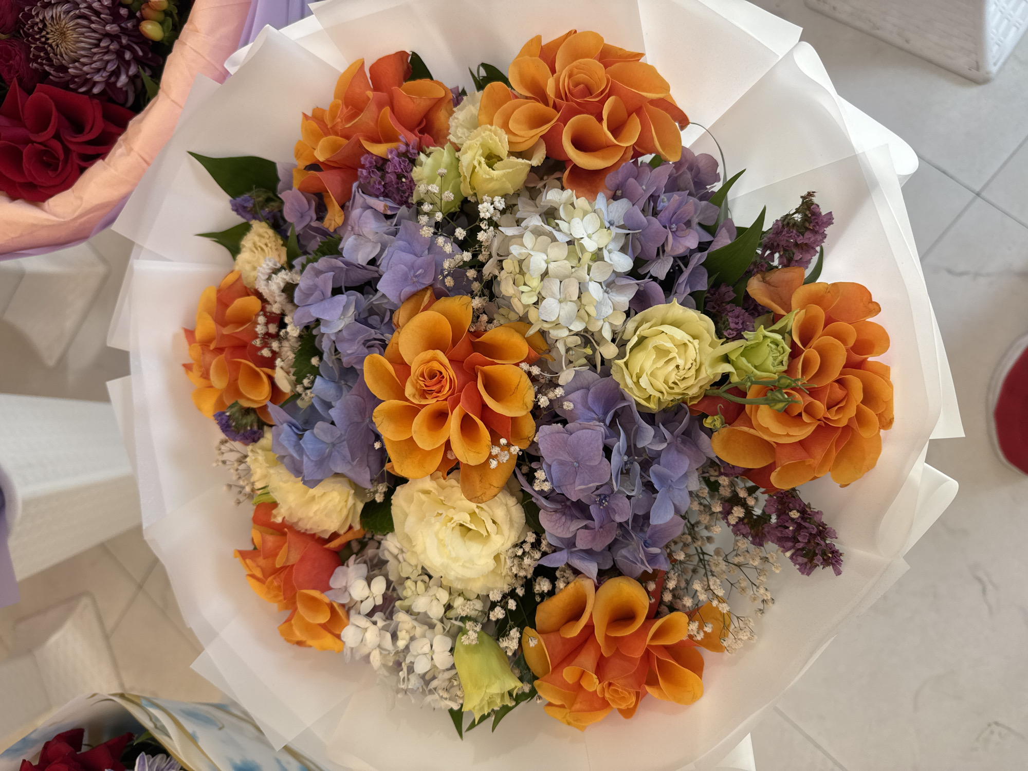 A top-down, medium shot captures a vibrant bouquet of flowers artfully arranged and wrapped in white paper. The bouquet is the primary subject, centrally positioned against a light-colored, possibly marble or tiled, surface. The flowers display a rich palette of colors, including bright orange, soft yellow, and varying shades of purple and blue, likely hydrangeas and roses. Small, delicate white flowers are interspersed throughout the arrangement, adding texture and volume. The white wrapping paper is translucent in places, with soft folds and curves. In the upper right corner, a portion of another bouquet, featuring dark red flowers and a textured purple bloom, is visible. The background is out of focus, with hints of a white structure or object to the left and right, possibly part of a floral display or a shop interior. No people or distinct actions are depicted. The scene appears to be an indoor setting, likely a flower shop. The location is not discernible from the visual information provided.