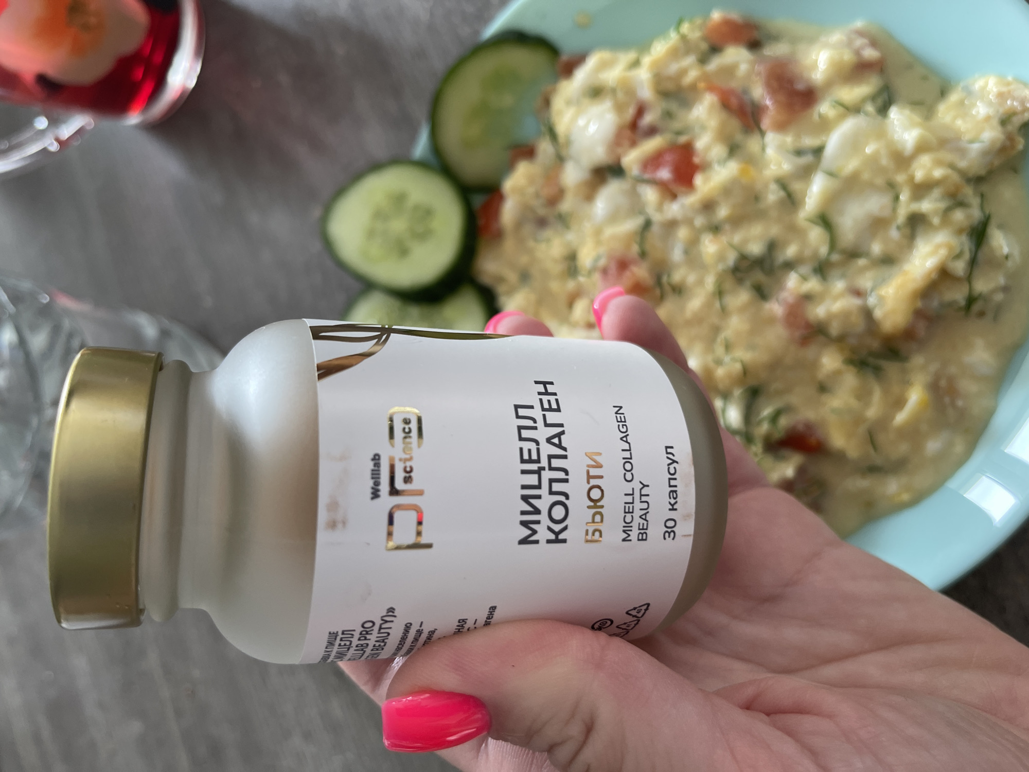 A close-up shot shows a person's hand with bright pink nail polish holding a white bottle of "MICELL COLLAGEN BEAUTY" supplements. The bottle has gold accents and Russian text. In the background, a plate of scrambled eggs with tomatoes and dill is visible, along with sliced cucumbers and a glass of water. The setting appears to be indoors. There is no information in the image to determine the location in Blagoveshchensk, Russia.