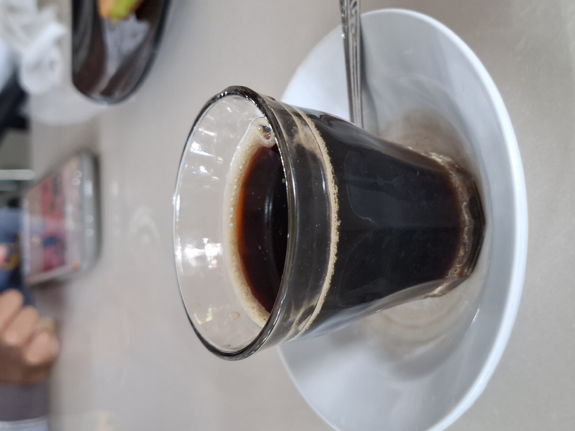 A glass of dark coffee rests on a white saucer with a spoon, atop a table. The scene is likely located in Lhokseumawe, Indonesia, but no specific details confirm the location. Other objects, including what appears to be another dish, a phone, and a person's hand are partially visible in the background.