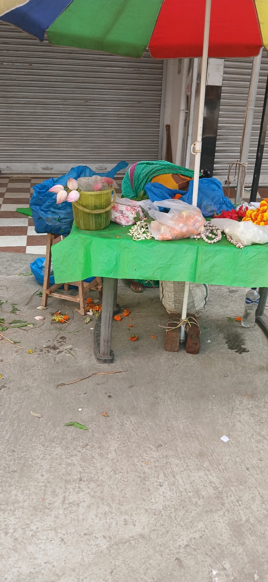 In Rajpur Sonarpur, India, an outdoor market stall is set up on a concrete surface in front of a closed metal roll-up shutter. A large, multi-colored umbrella, segmented into red, green, and blue, provides shade over the stall. The stall itself is a green plastic-covered table supported by a wooden stool on the left and a metal leg on the right. On the table are various items, including plastic bags filled with flowers, a yellow bucket containing pink lotus flowers, and a pile of green fabric. A person, only their feet and lower legs visible, is seated behind the table, wearing sandals. Scattered on the concrete floor around the stall are fallen flower petals and leaves, along with a water bottle and two bricks used to stabilize one of the table legs. The overall scene depicts a vendor preparing to sell flowers or related items.