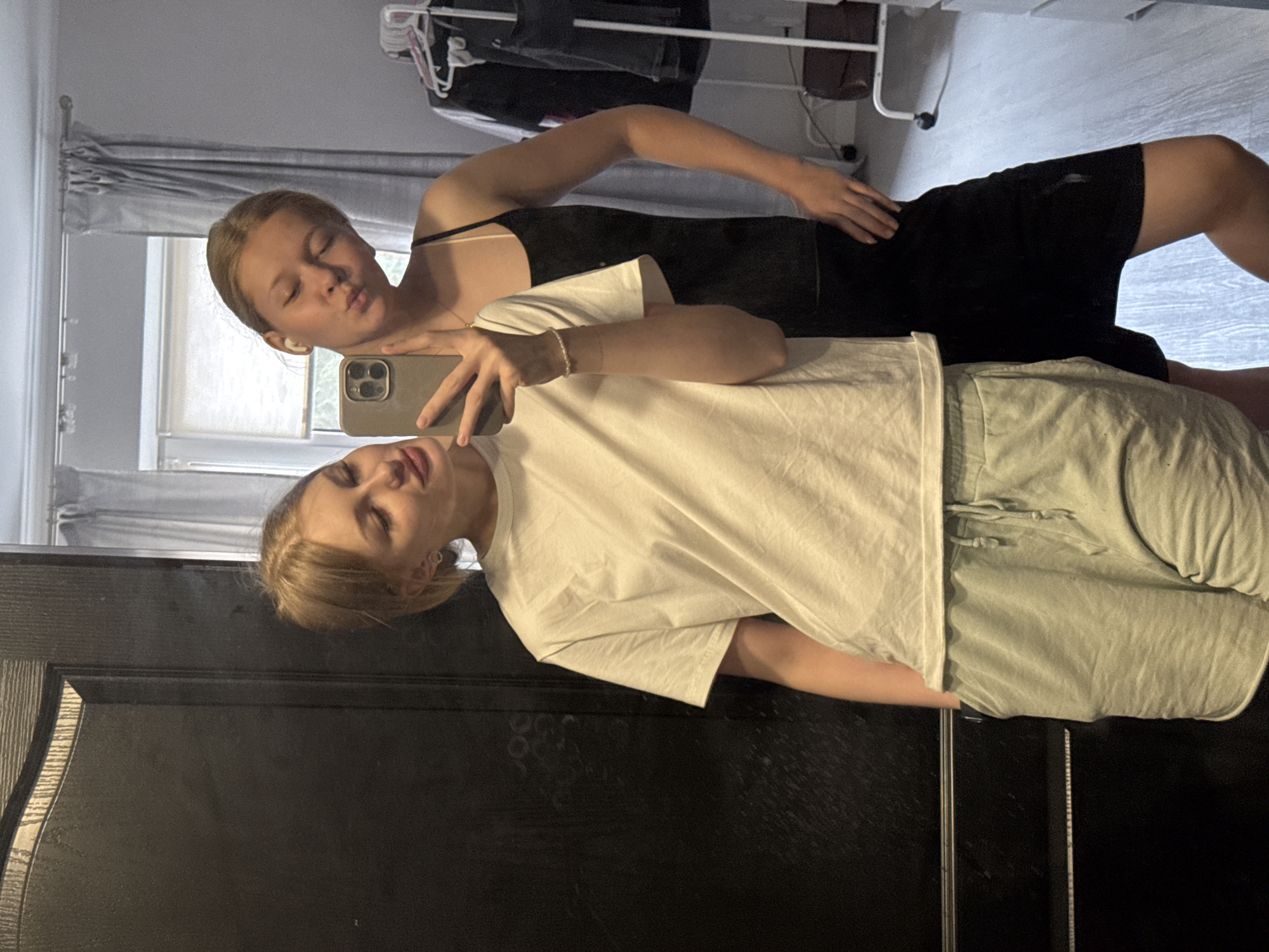 This media file features two young women posing for a mirror selfie in what appears to be a domestic setting. The image is captured in portrait orientation.
The foreground is dominated by a dark, reflective surface, likely a mirror, which shows the two subjects. The woman closer to the viewer is wearing a light-colored t-shirt and light green shorts. She is holding a smartphone in her left hand, angled to take the picture, and looking towards the camera with a neutral expression. A delicate bracelet adorns her wrist.
Behind her, a second woman is visible, also reflected in the mirror. She is wearing a black outfit, possibly a dress or top and shorts. She is leaning forward, with her eyes closed and lips puckered as if blowing a kiss, and her right hand rests on her hip. Her left hand is also holding a smartphone. Both women have light-colored hair.
The background, reflected in the mirror, provides some context about the environment. There are grey curtains covering a window, and above them, a clothes rack with some garments hanging. The floor appears to be a light gray laminate or tile. The overall impression is a casual, indoor scene, likely a bedroom or dressing area. The lighting suggests it could be daytime, but it is difficult to determine the exact time of day or weather conditions outside. The mood seems playful and relaxed. There is no visible text in the image.